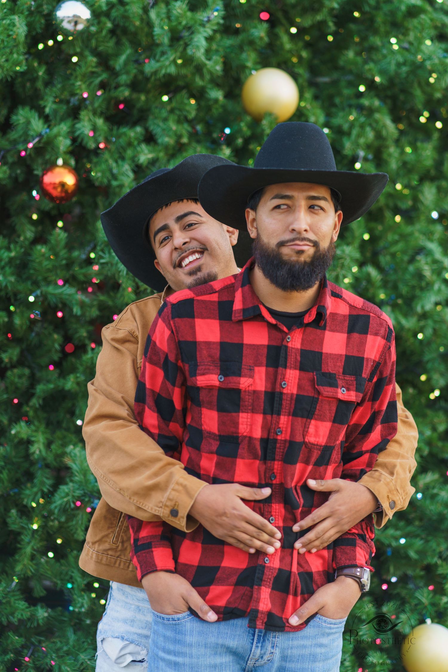 Two brothers wearing cowboy hats standing together in front of a decorated Christmas tree, smiling during a holiday portrait.
