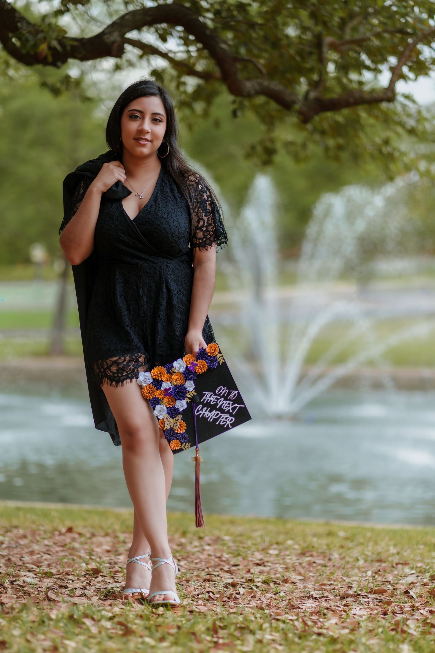 A young woman in a black lace dress holds a decorated graduation cap near a fountain outdoors.