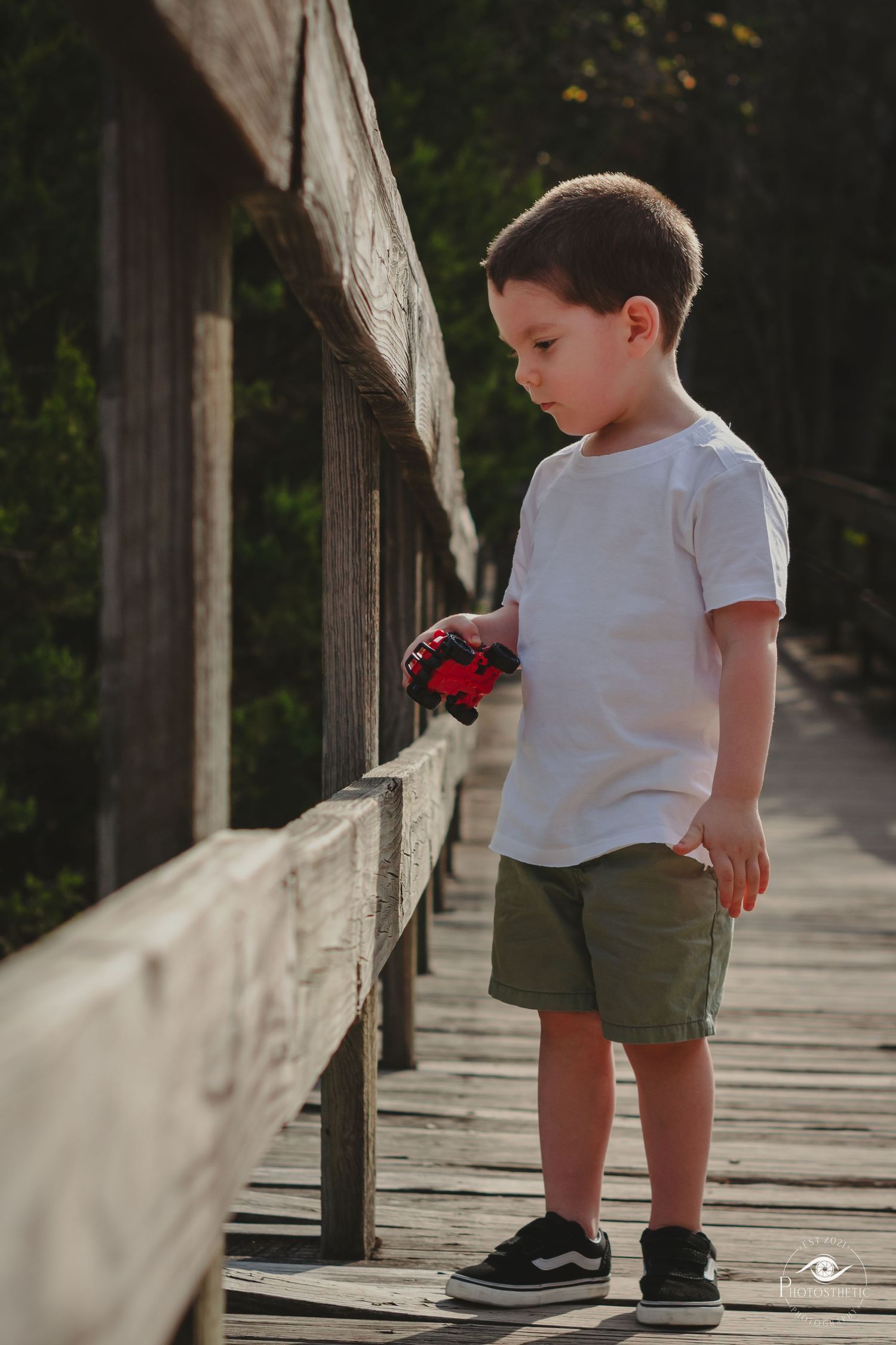 A small child in a white t-shirt and olive shorts explores a wooden boardwalk on a sunny day.