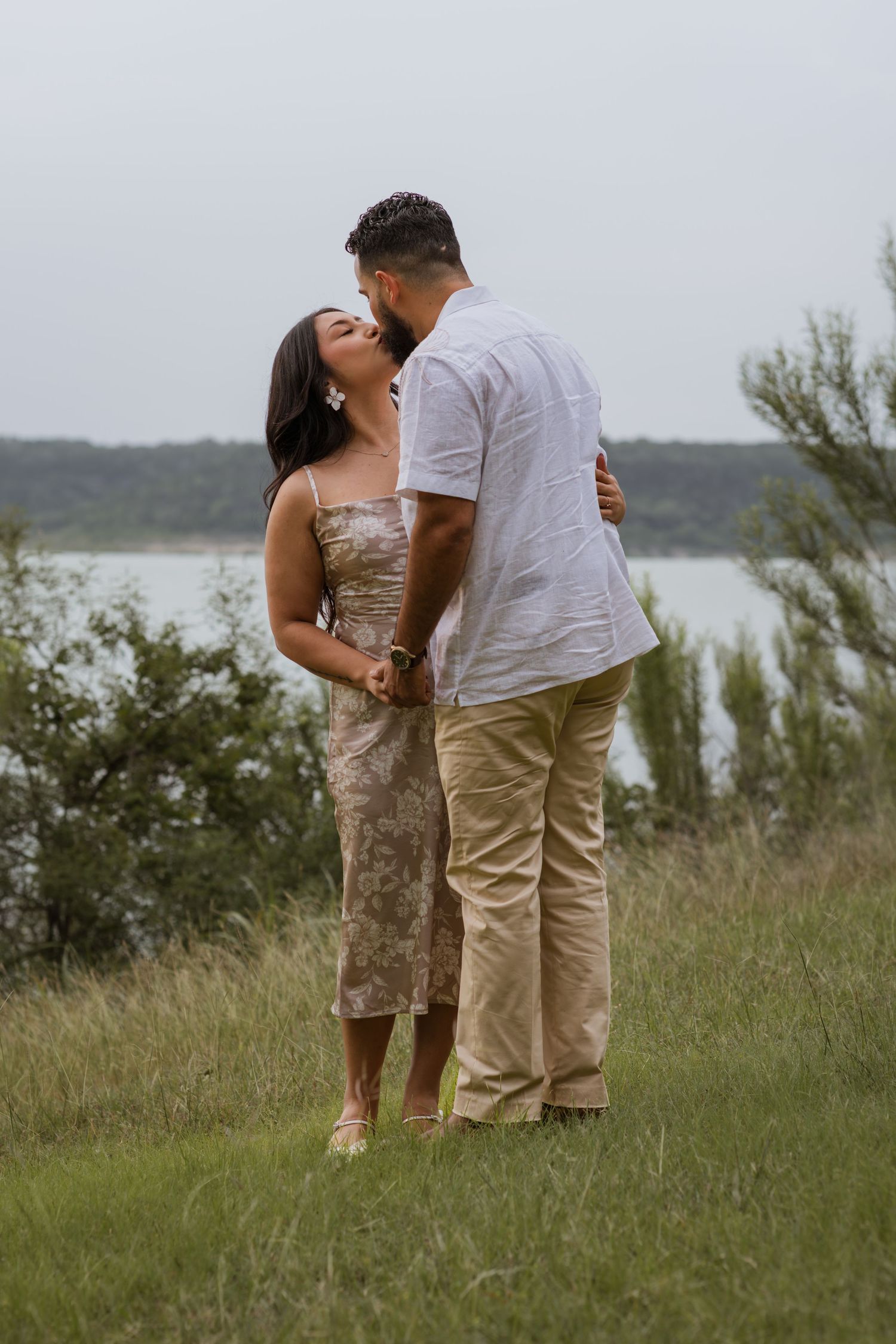 Couple sharing a romantic kiss outdoors near a lake, woman in floral midi dress, man in white shirt and khaki pants.
