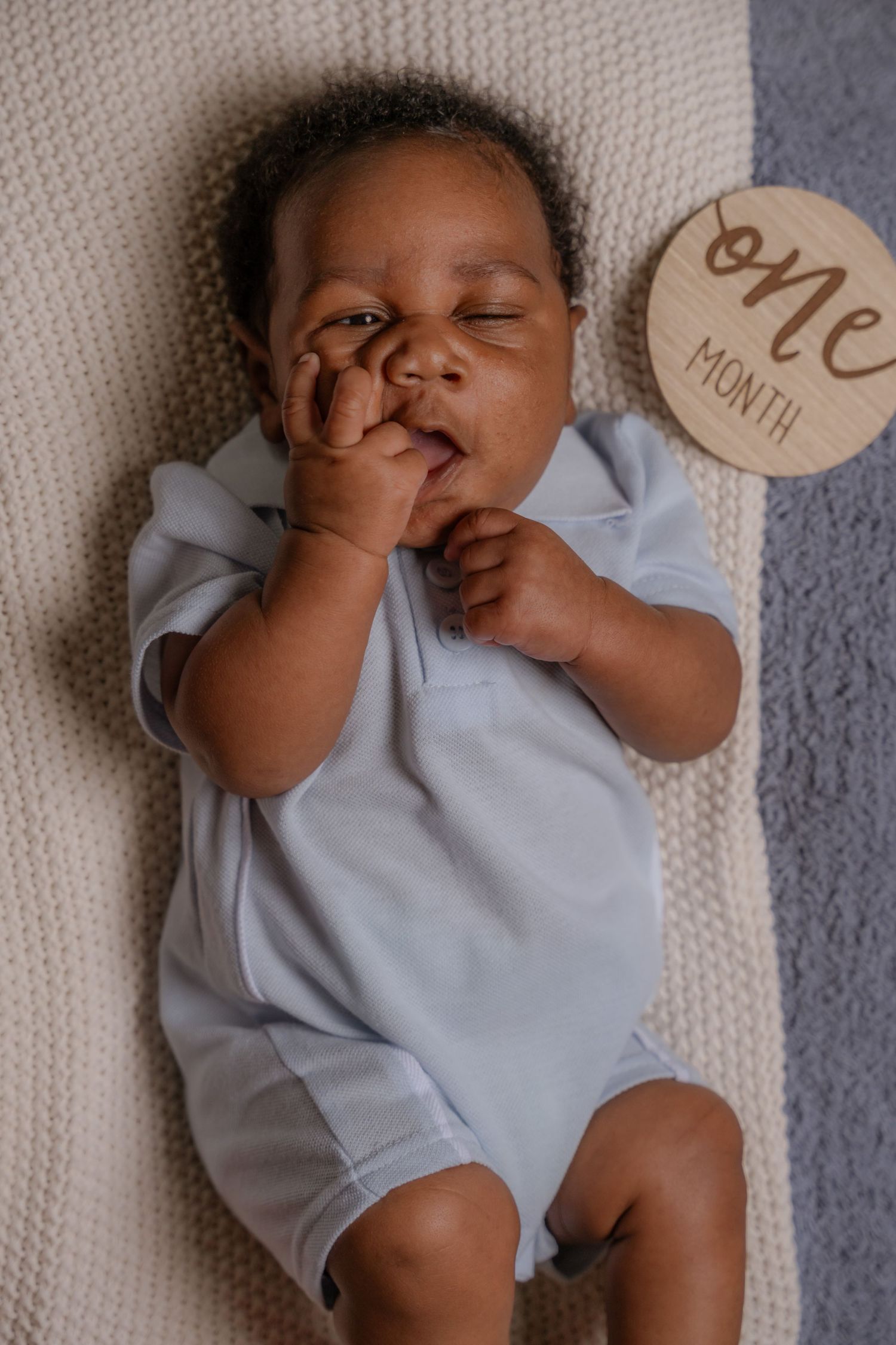 One month old baby in light blue onesie lying on knit blanket with wooden milestone disc.