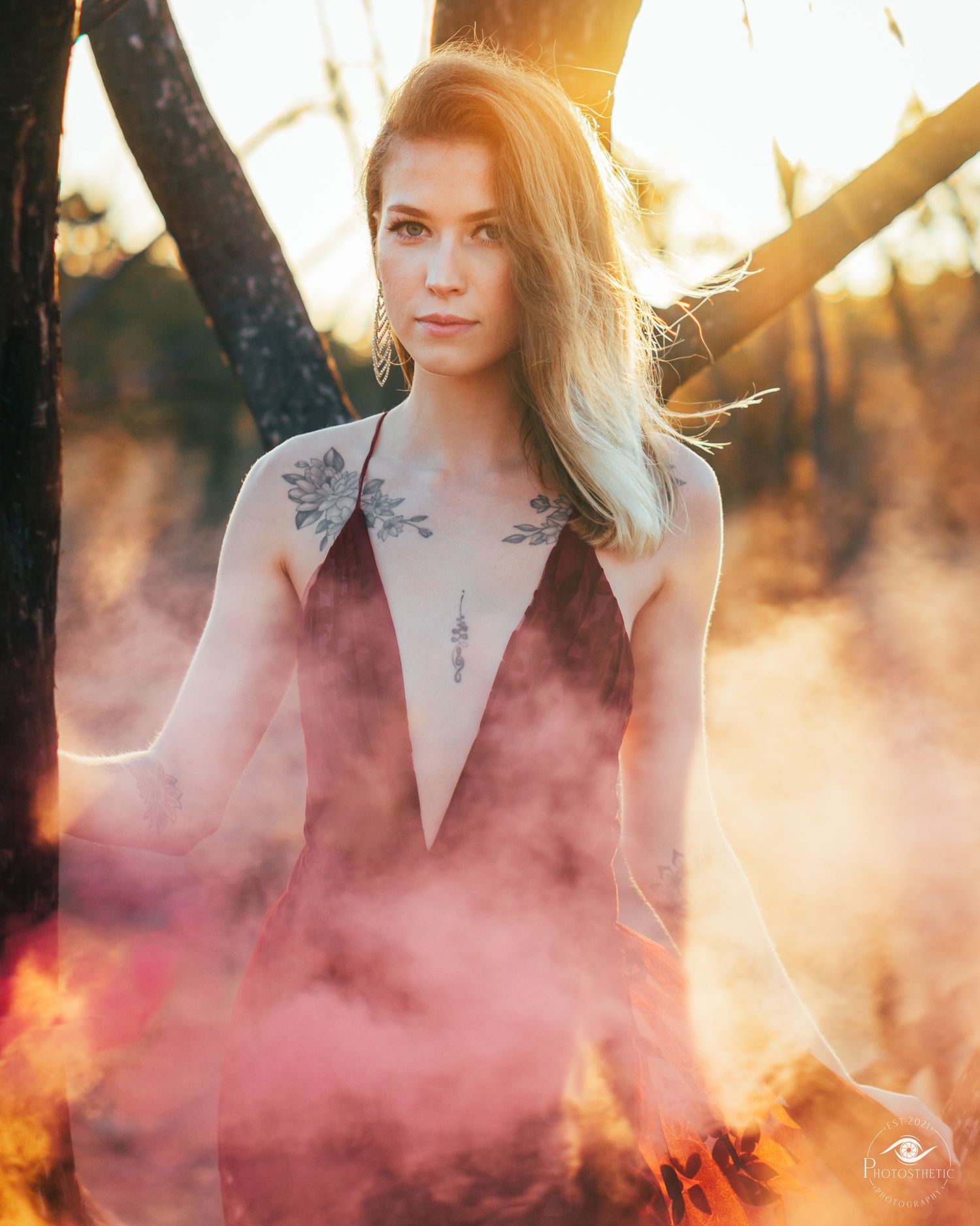 Woman with tattoos wearing a deep V-neck dress standing outdoors at golden hour with soft colored smoke.