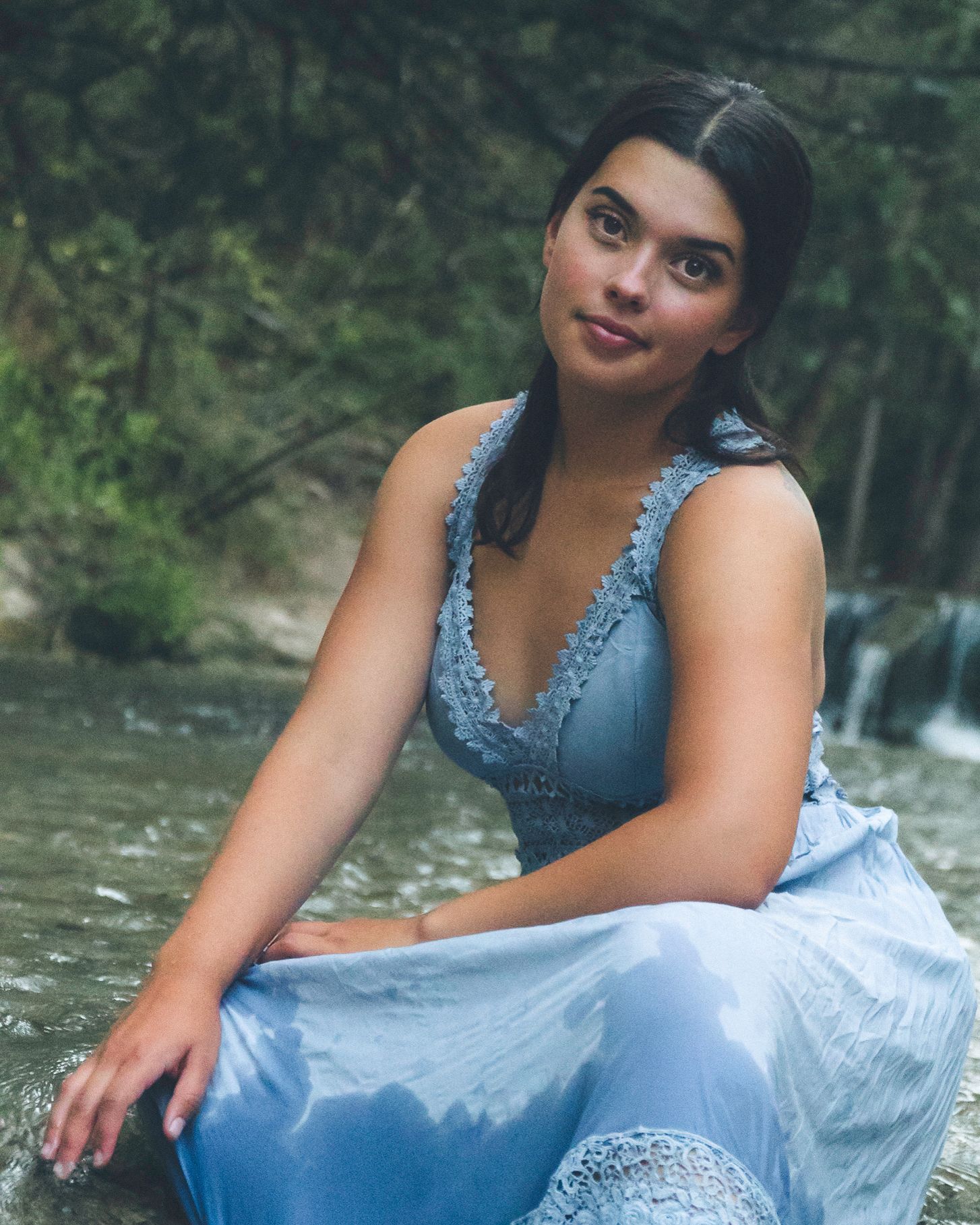 Woman wearing a light blue dress sitting beside a creek surrounded by greenery during an outdoor portrait session.