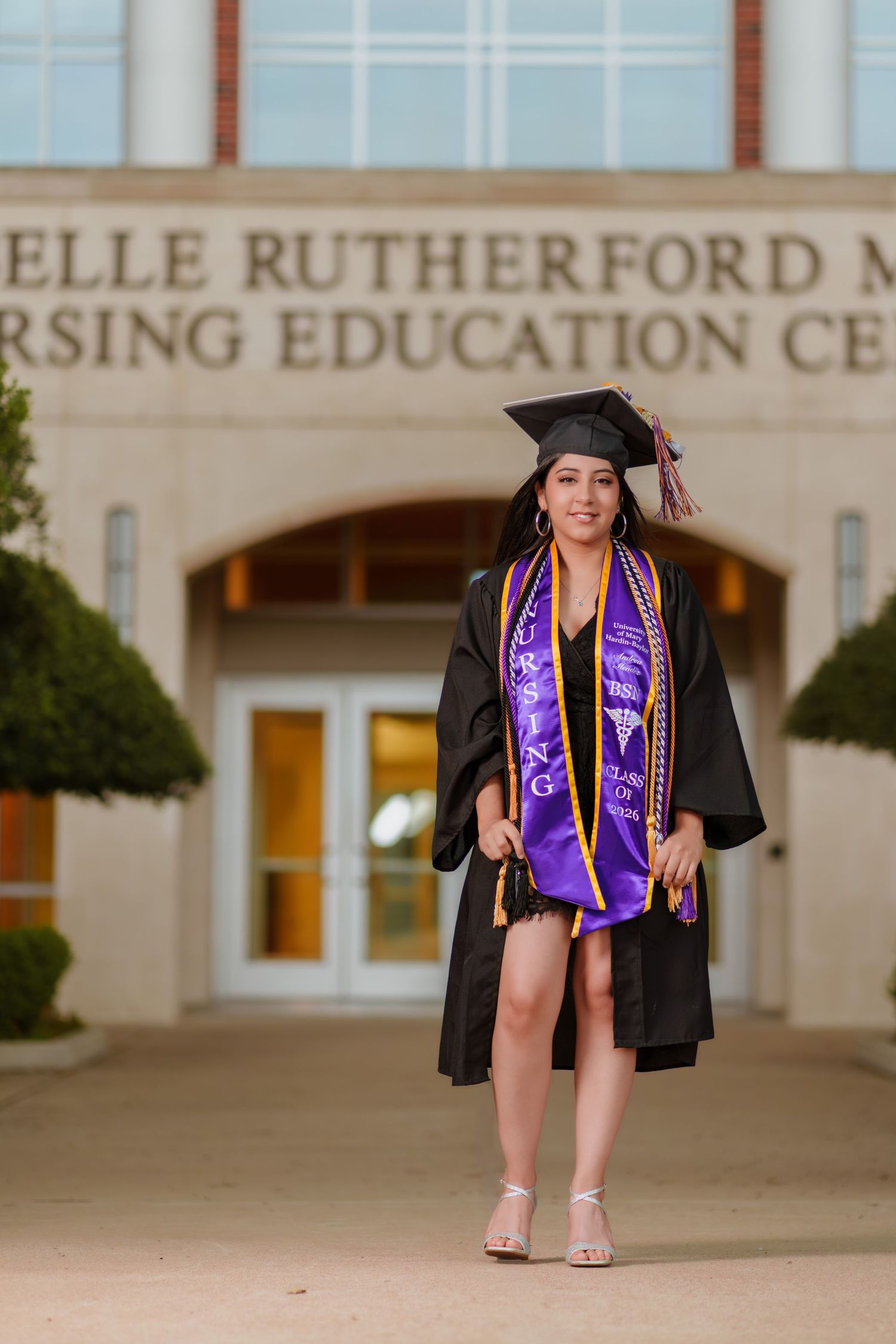 Female graduate in black cap and gown with purple stole poses in front of Nursing Education Center building.