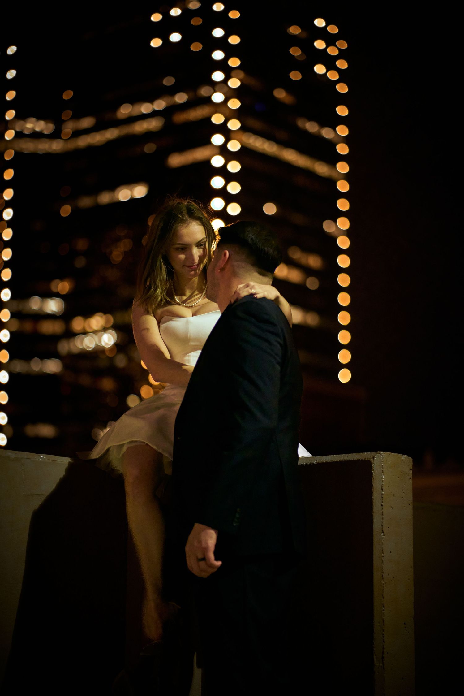 Romantic couple poses against backdrop of twinkling lights in evening setting.