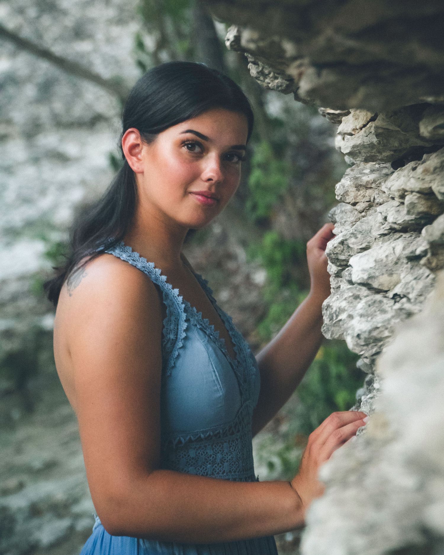 Outdoor portrait of a woman in a blue dress touching a textured rock wall in natural light.