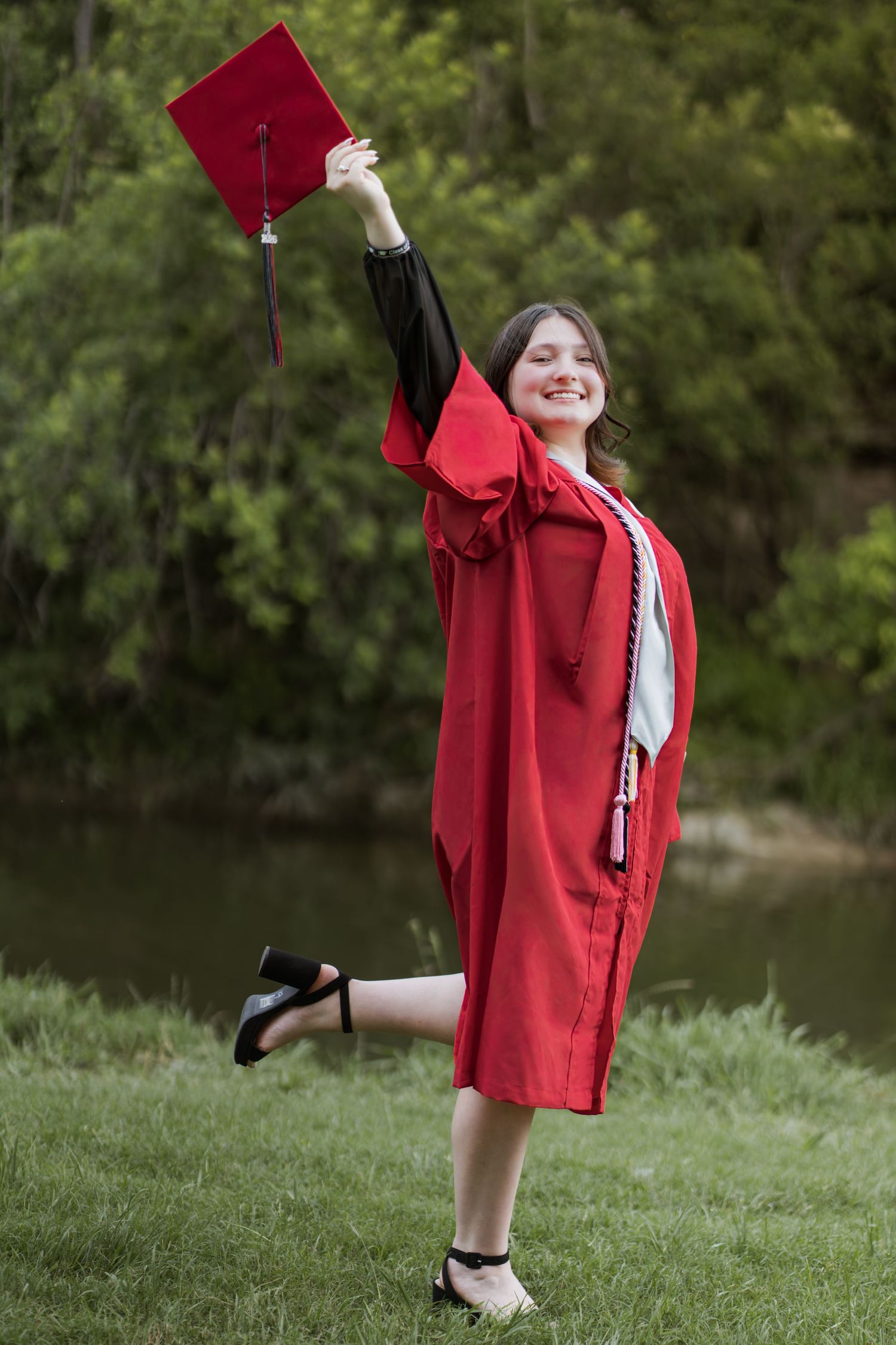 Young woman in red graduation gown and cap celebrates outdoors, raising her mortarboard triumphantly while standing on one leg.