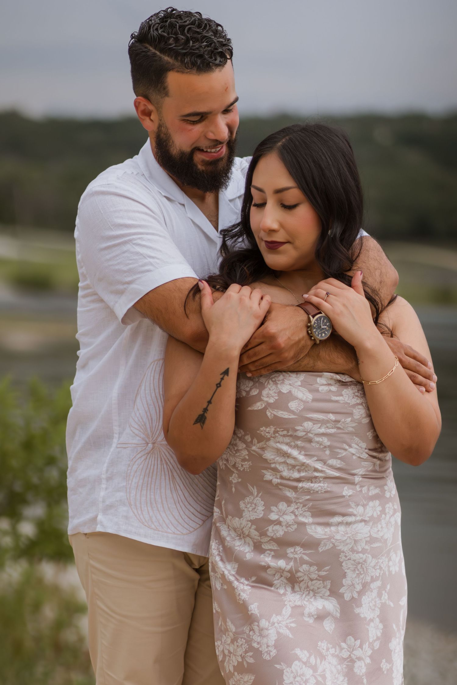 A bearded man in a white shirt embraces a woman in a floral dress outdoors, both smiling softly in a romantic pose.