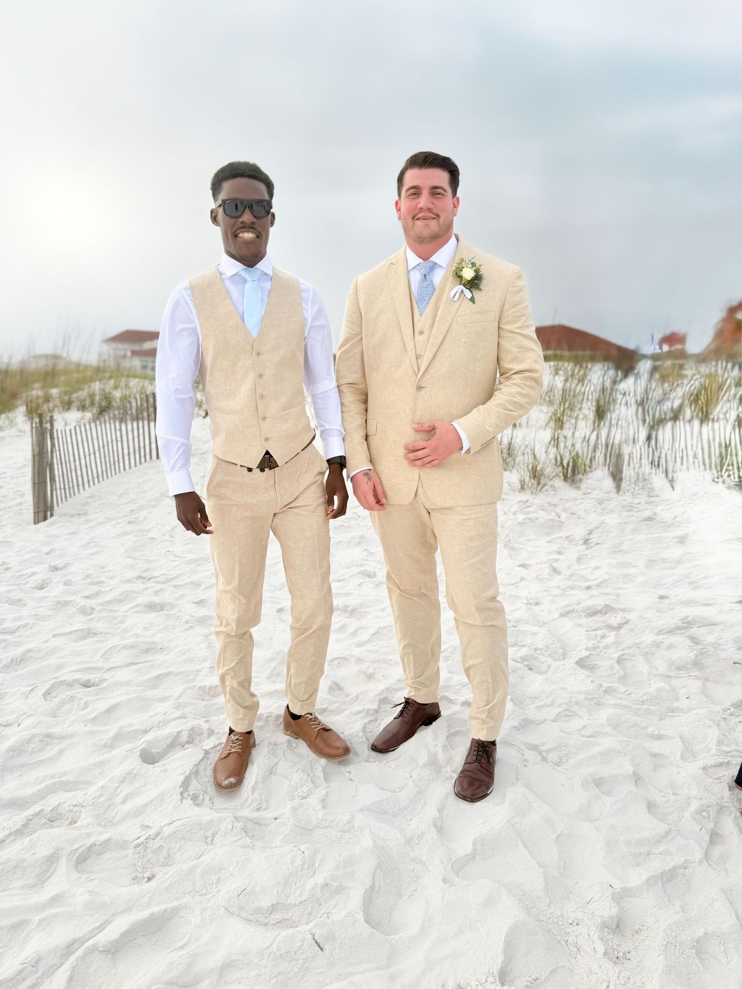 Wedding party members in tan suits pose together on white sand beach.