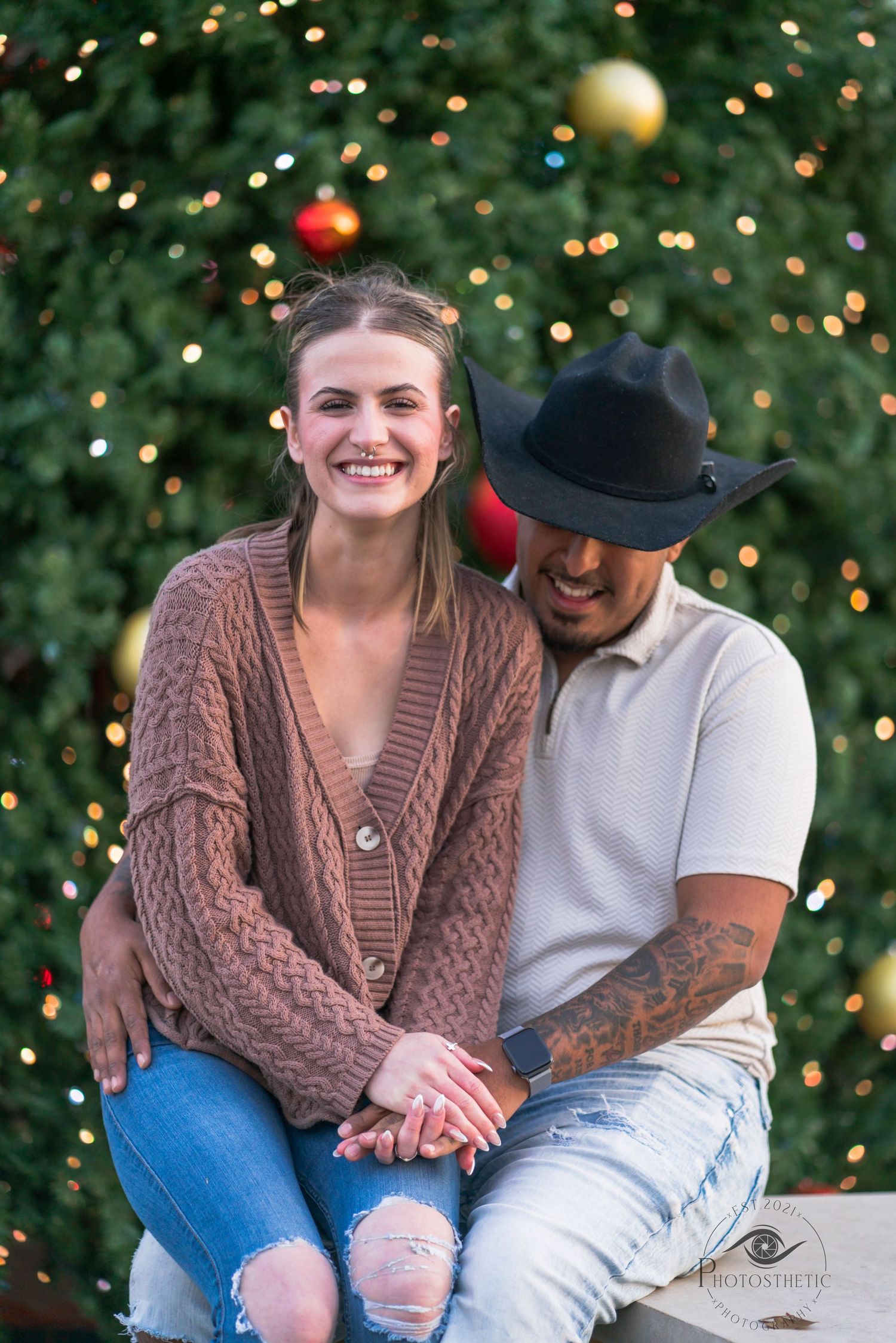 Couple sitting together outdoors in front of a decorated Christmas tree, smiling during a holiday portrait.
