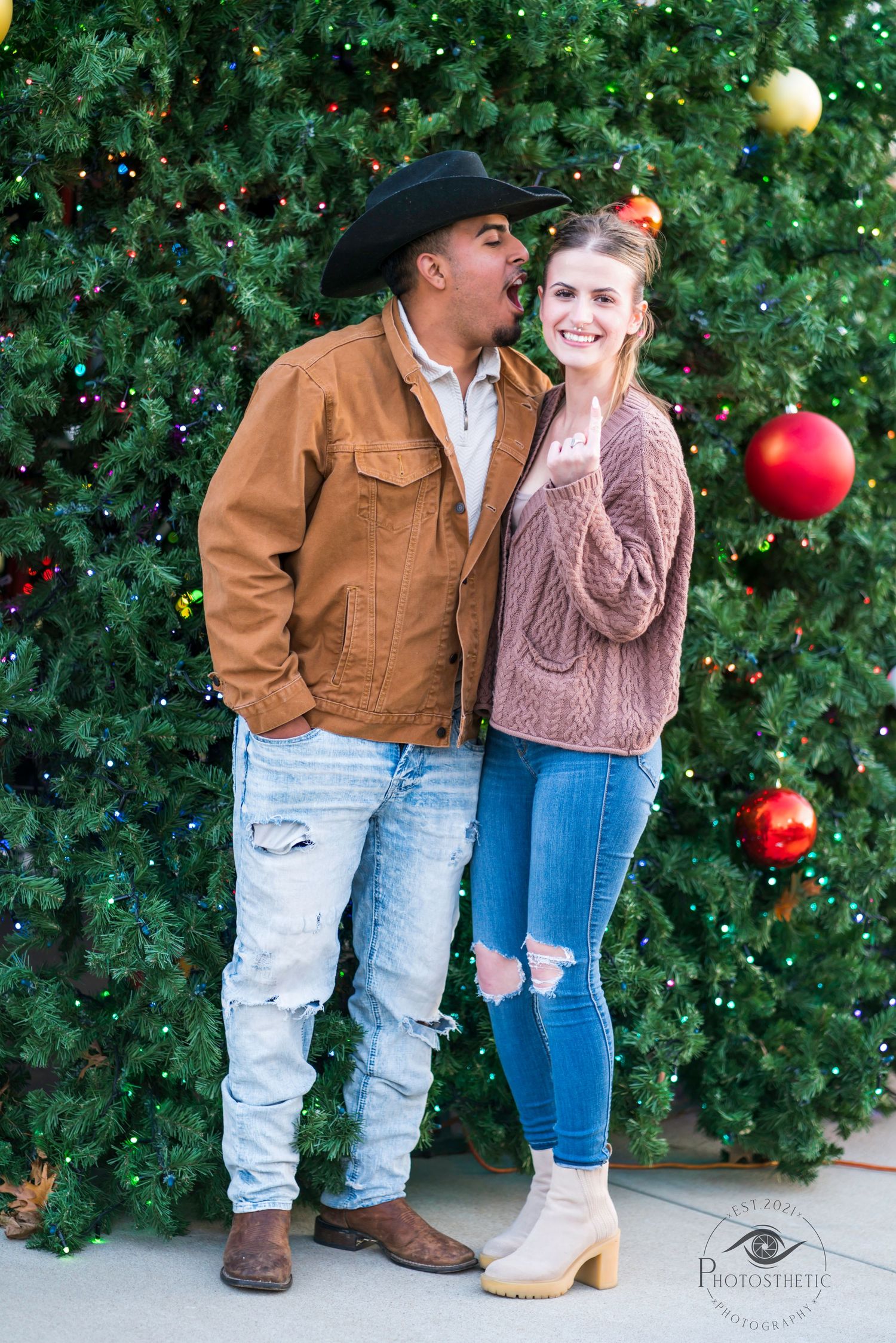 A couple poses together in front of a decorated Christmas tree with large red ornaments.
