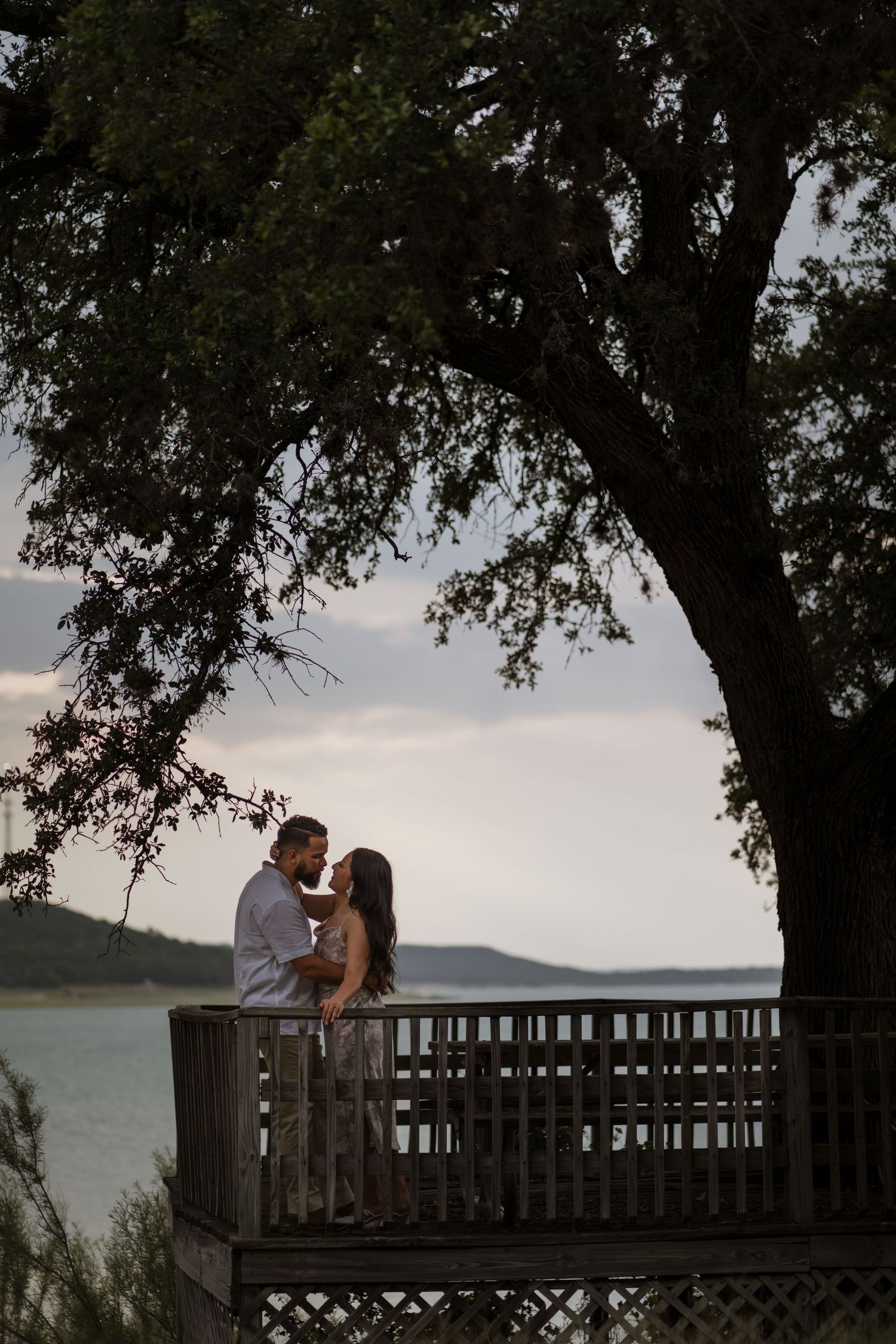 Couple sharing a romantic moment on a wooden deck overlooking a serene lake, framed by a large tree at dusk.