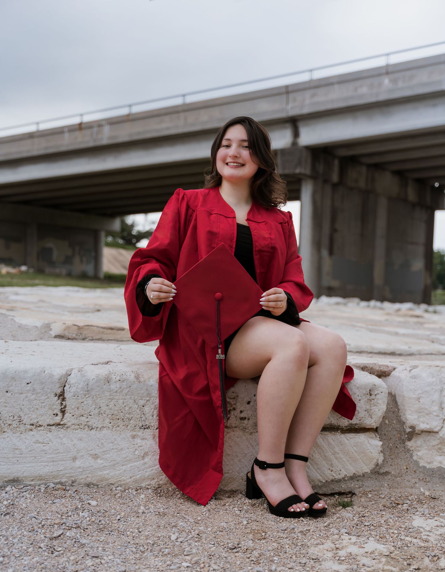 Young woman in red graduation cap and gown sitting on stone steps outdoors, smiling proudly on her graduation day.