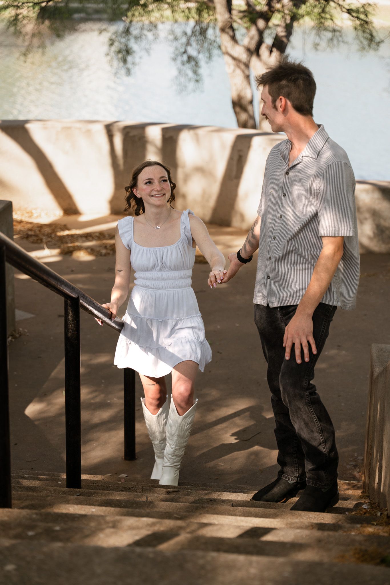 Couple holding hands walking up outdoor steps, woman in white dress and cowboy boots, man in gray shirt and dark jeans.