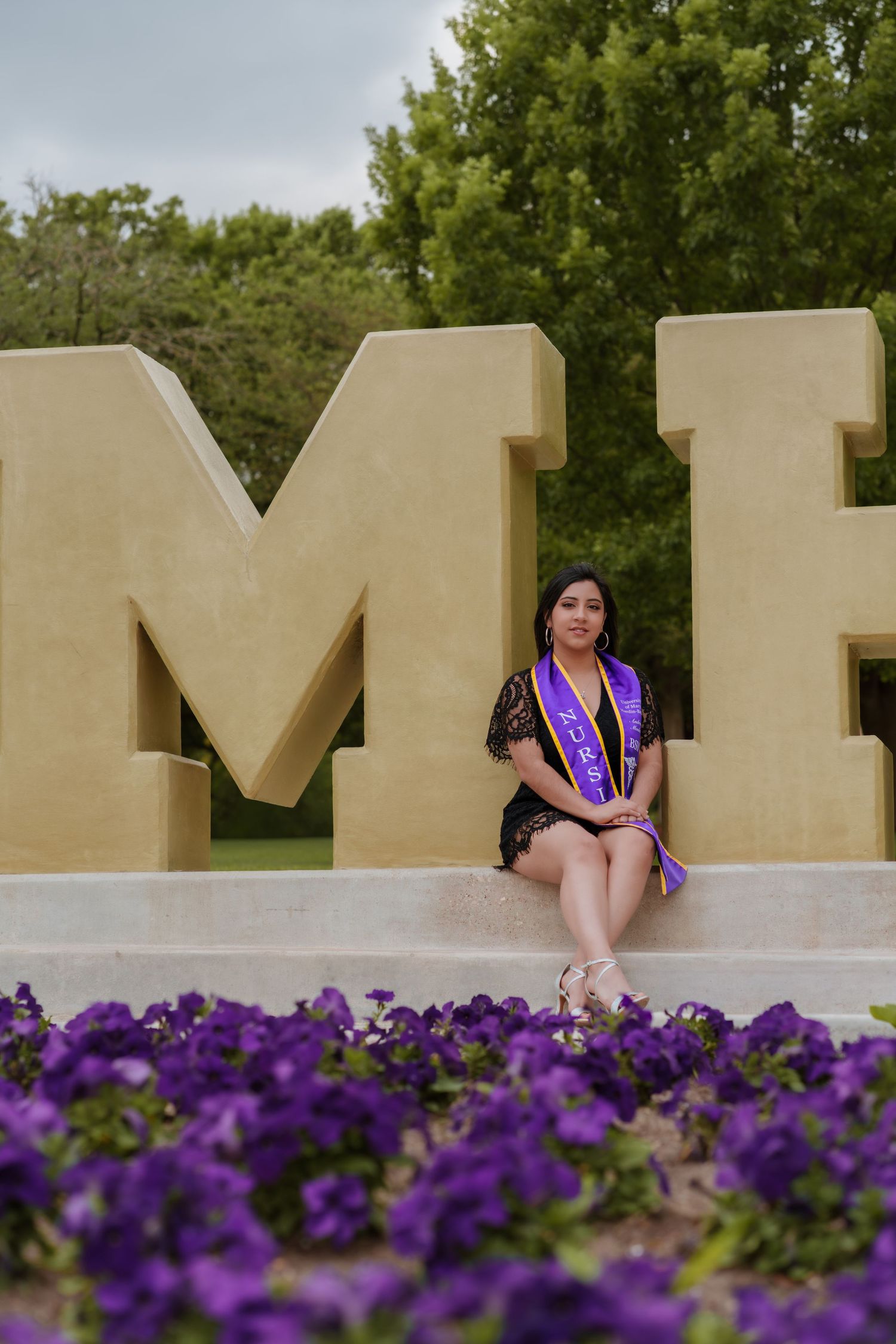Graduate in black dress and purple sash poses by large university letters with purple flowers in foreground.
