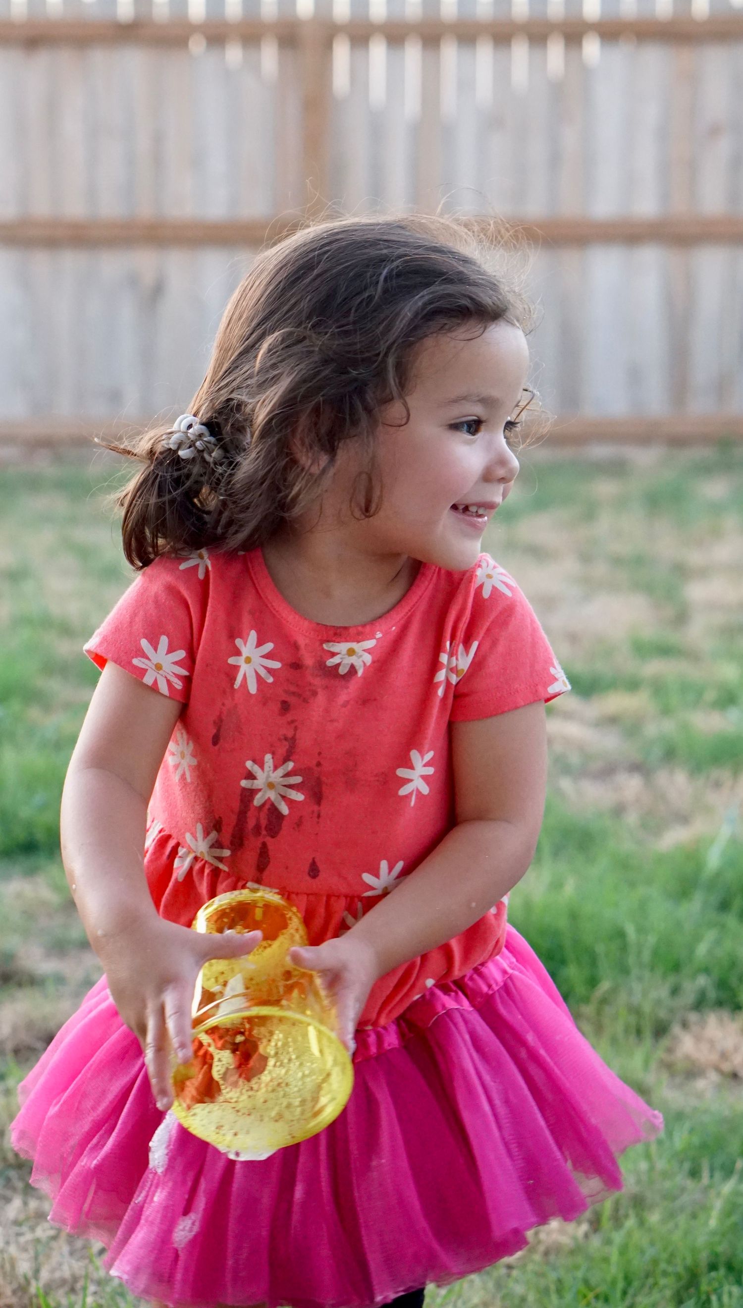 Child in pink tutu dress smiles while holding yellow toy outdoors.