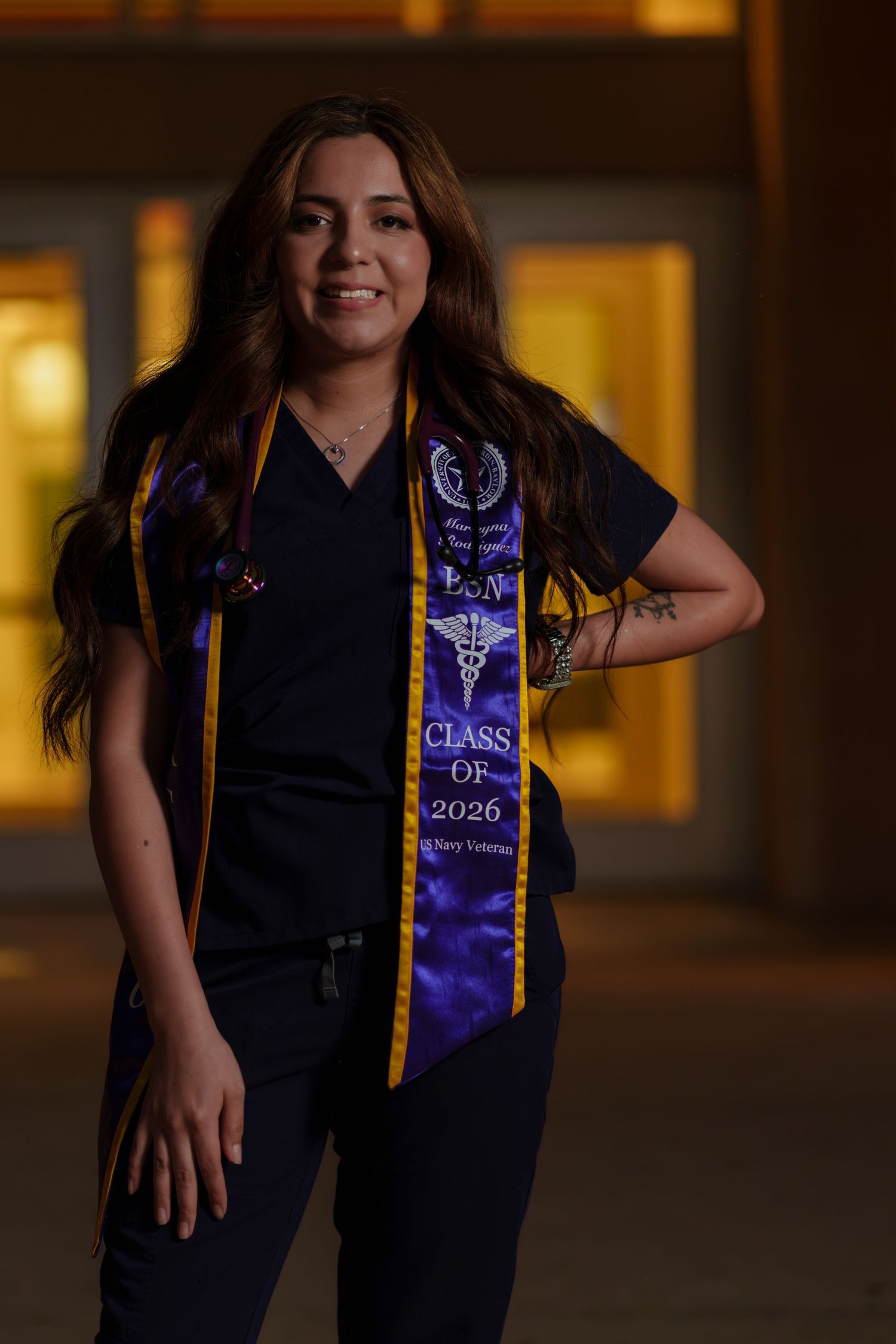Nursing student in scrubs wearing a purple and gold Class of 2026 graduation stole with caduceus symbol posing at night.