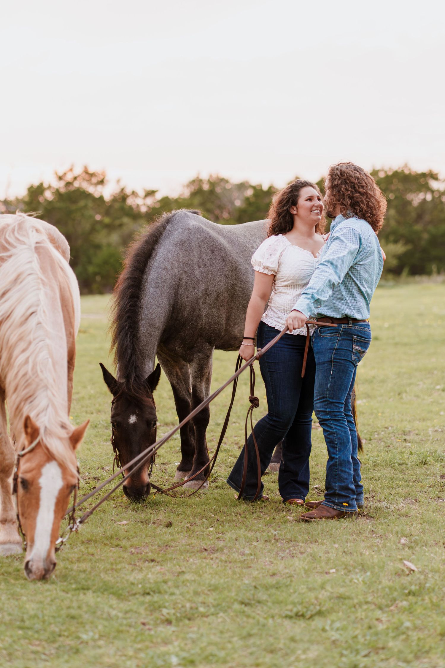 Two women laughing together while holding horses on a green pasture at sunset.