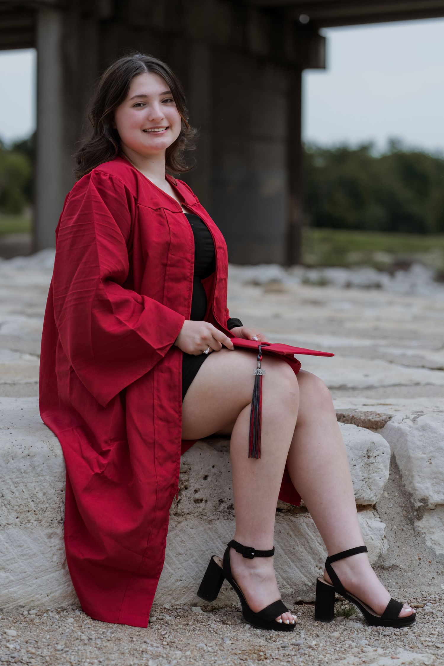 Smiling graduate in red cap and gown sitting on stone steps outdoors holding graduation cap with tassel.