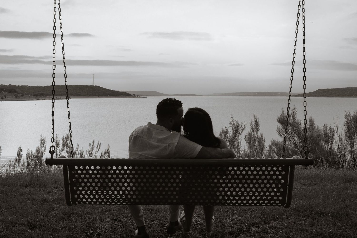 Black and white photo of a couple sitting on a swing bench overlooking a calm lake at dusk.