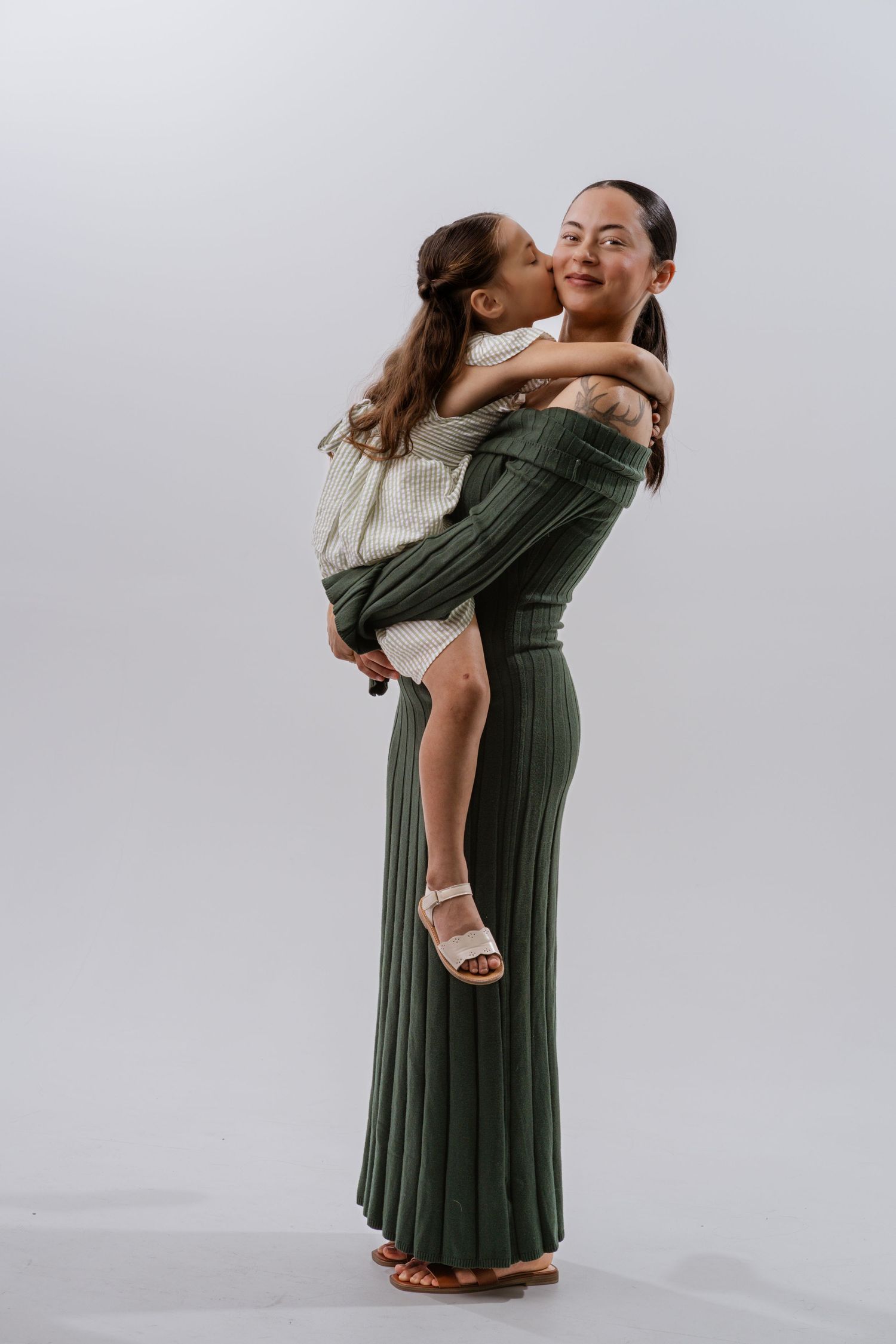 A woman in green wide-leg pants holds a young girl who kisses her cheek, both smiling on a white background.