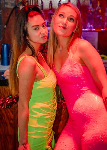 Two people in bright neon outfits pose together under red lighting at a nightclub or party venue.