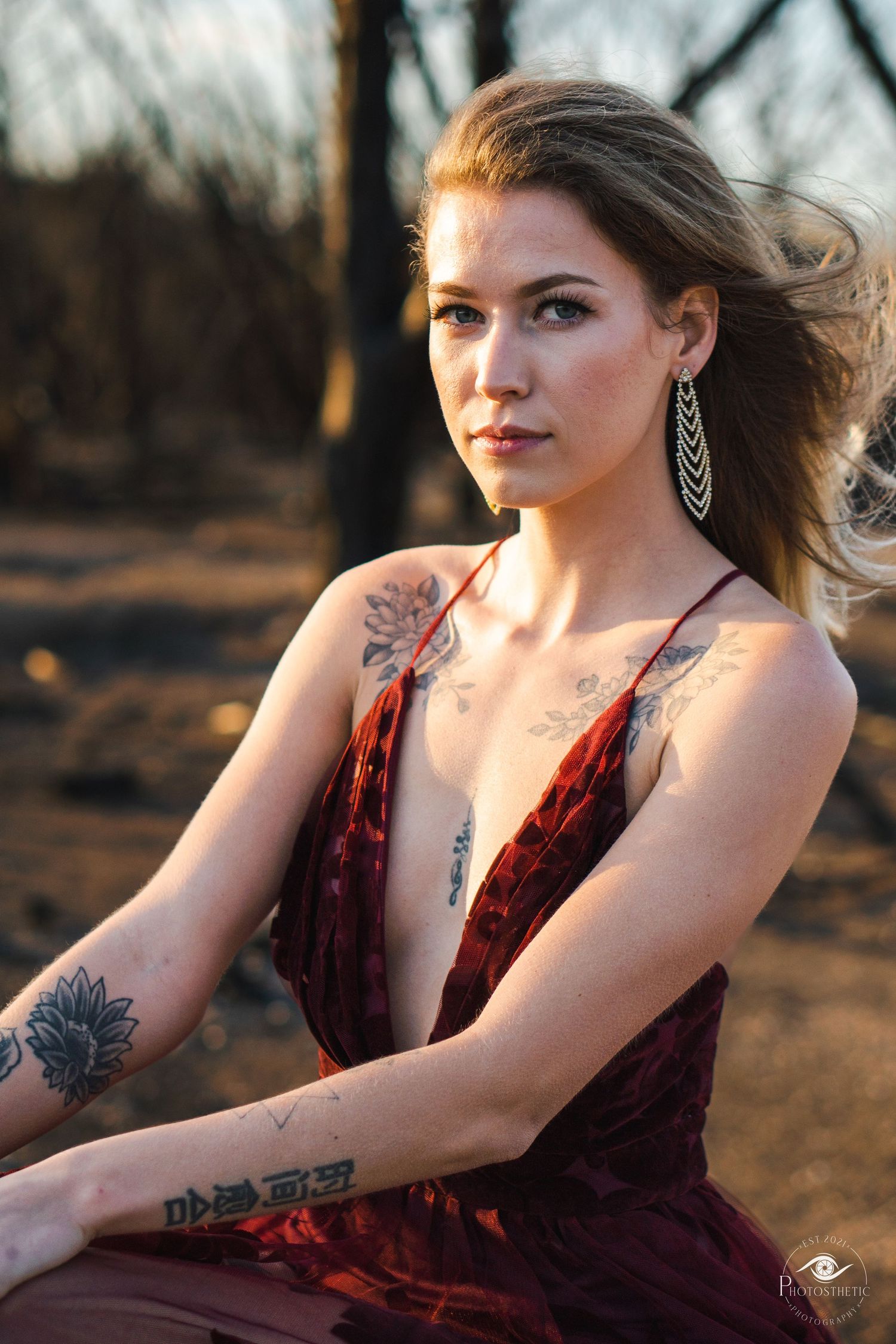 Outdoor portrait of a woman with tattoos wearing a deep red dress, seated in natural light.