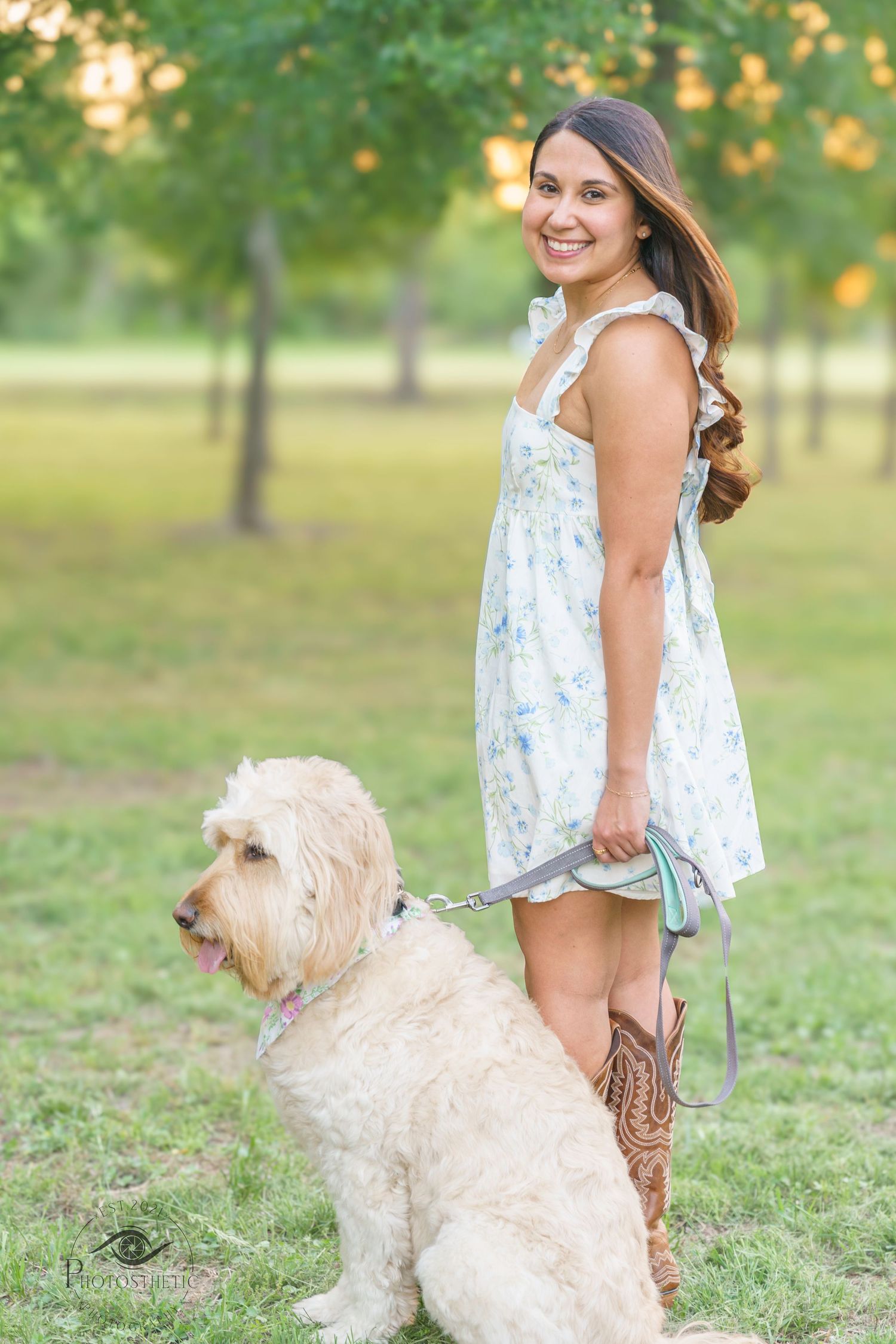 Woman in a light floral dress standing in a park holding a leash with her dog during golden hour.
