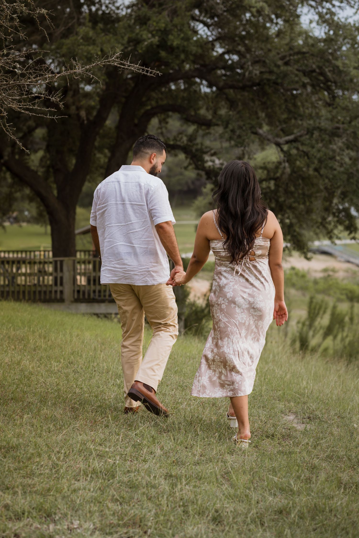 A couple holds hands while walking through a lush green field, the woman in a floral dress and man in white shirt.