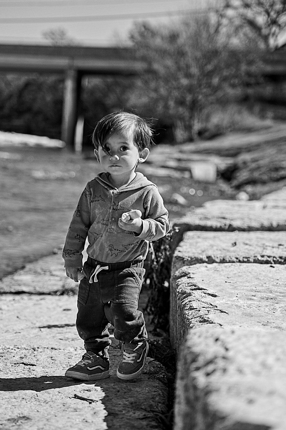 Black and white portrait of a toddler standing on a stone path near a creek, holding a small object.