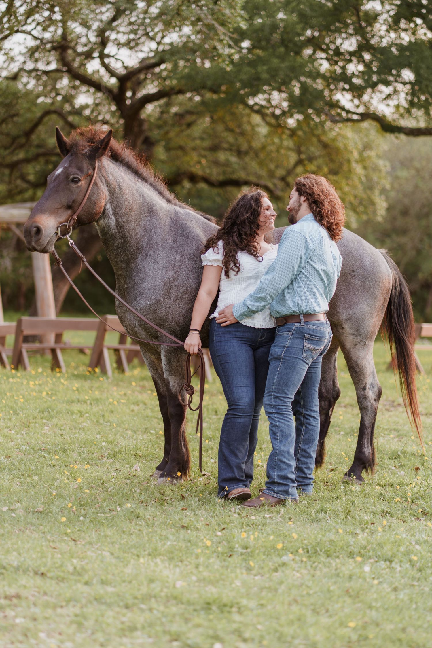 A couple stands together beside a gray horse in a green pasture, sharing a romantic moment outdoors.