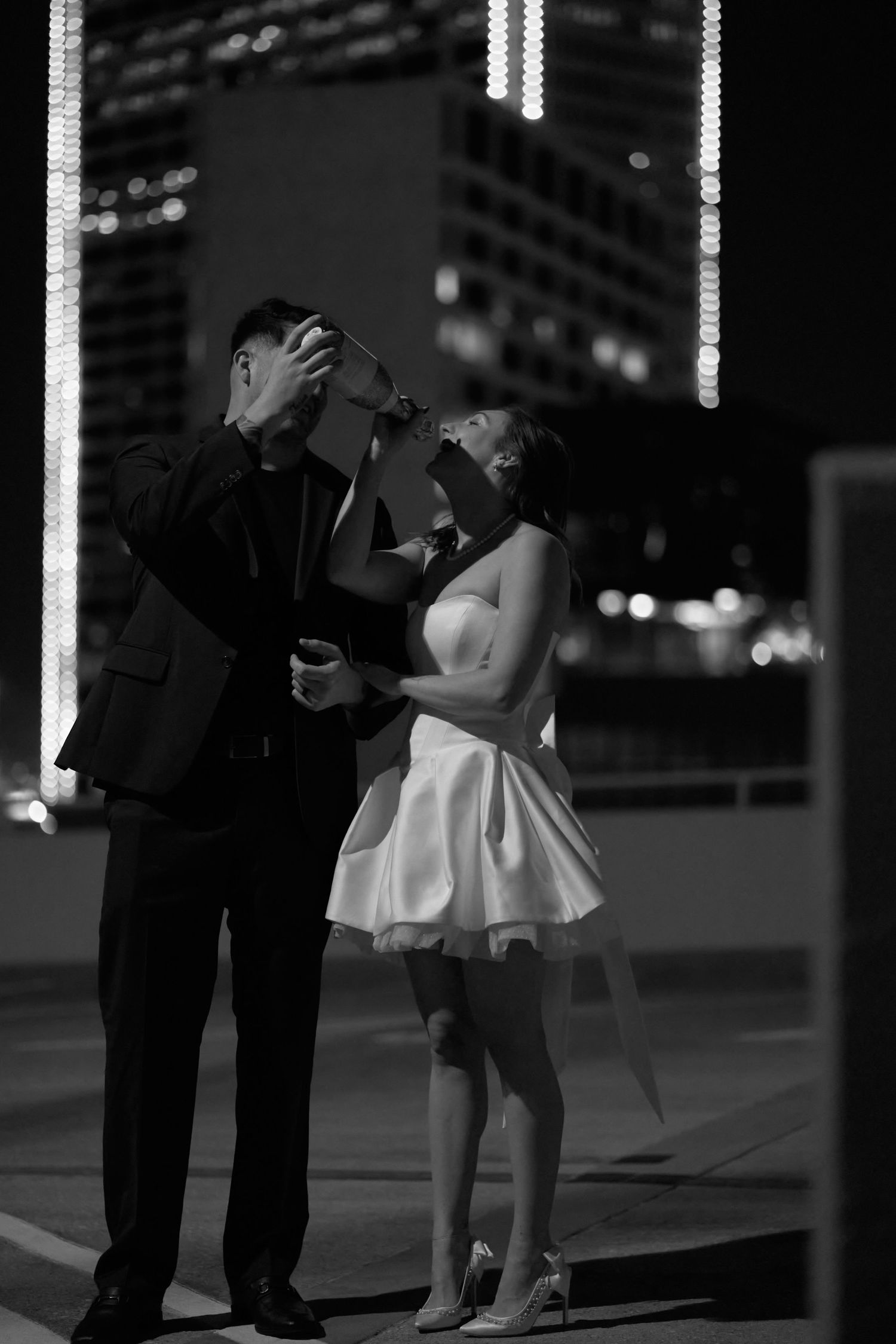 A romantic black and white nighttime scene showing a couple in formal attire sharing a moment on a city street.