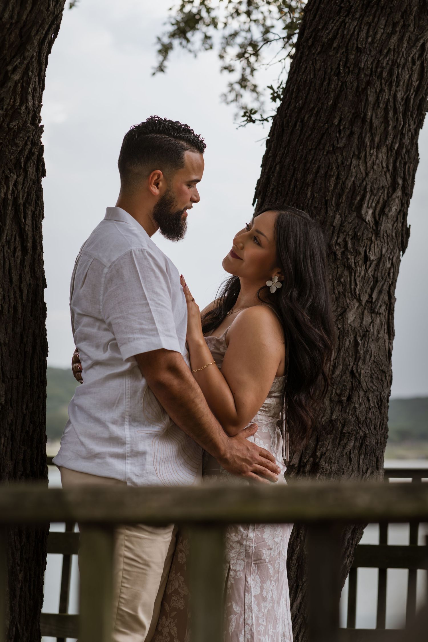 Romantic couple embracing between tree trunks near a wooden railing, gazing into each other's eyes.