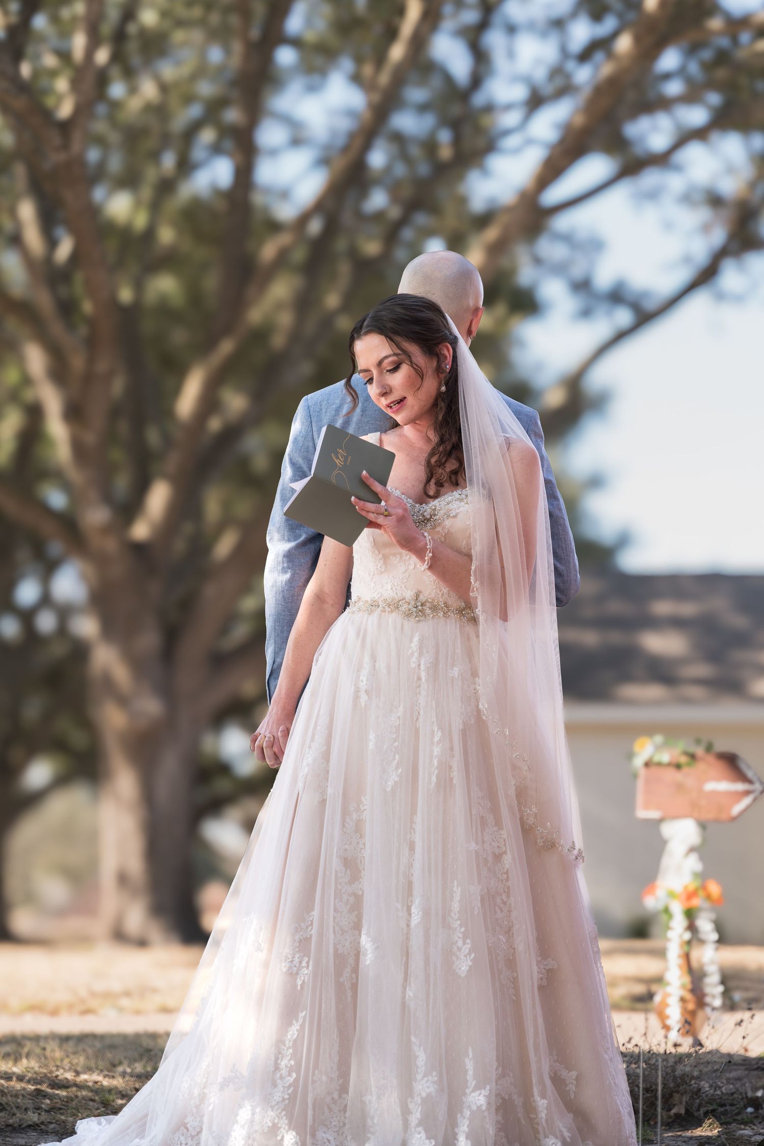 Bride standing outdoors in a lace wedding gown holding a vow book during a ceremony.