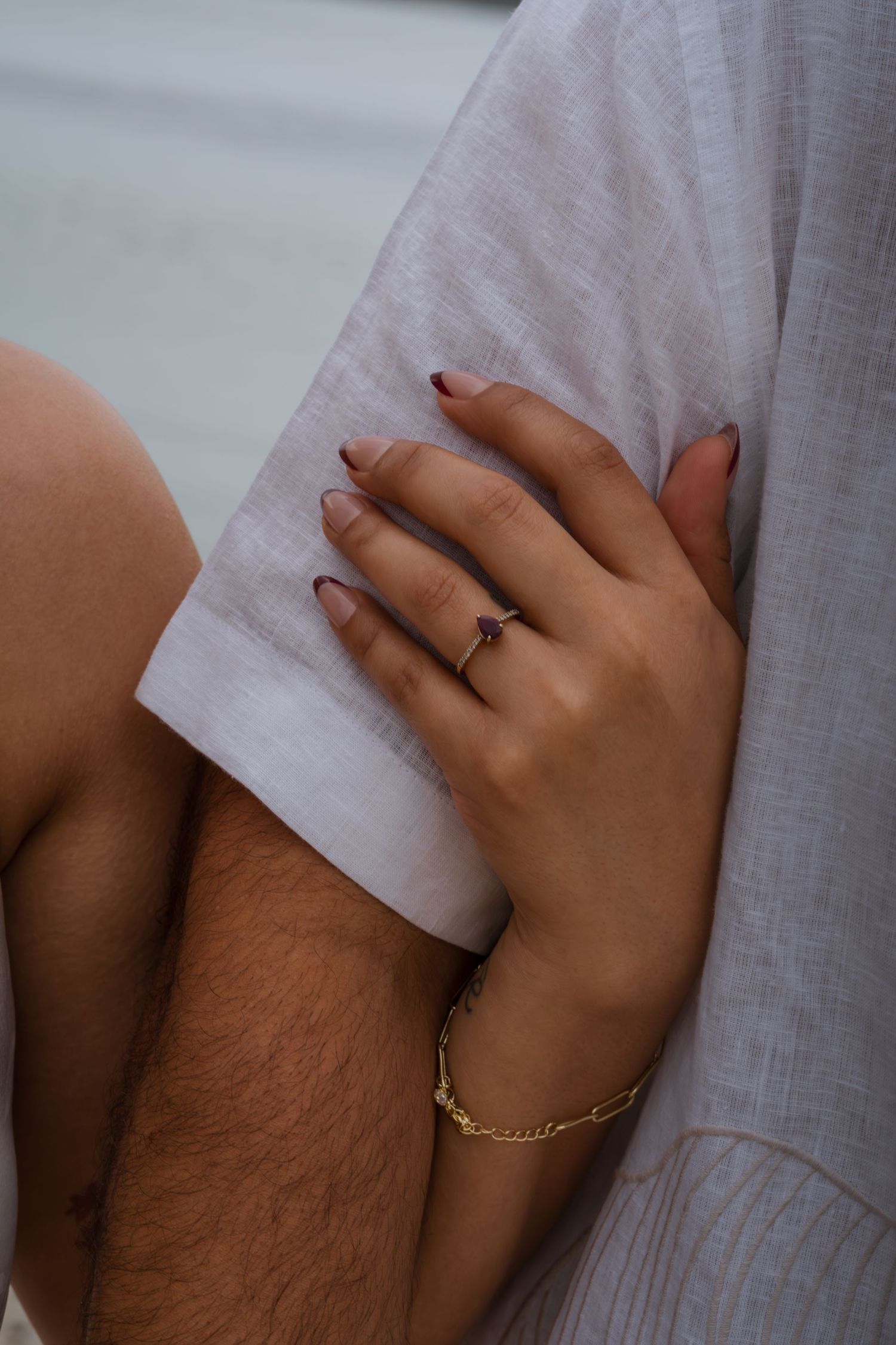 Close-up of a woman's hand with dark nails and gold bracelet, wearing an engagement ring, resting on a man's arm.