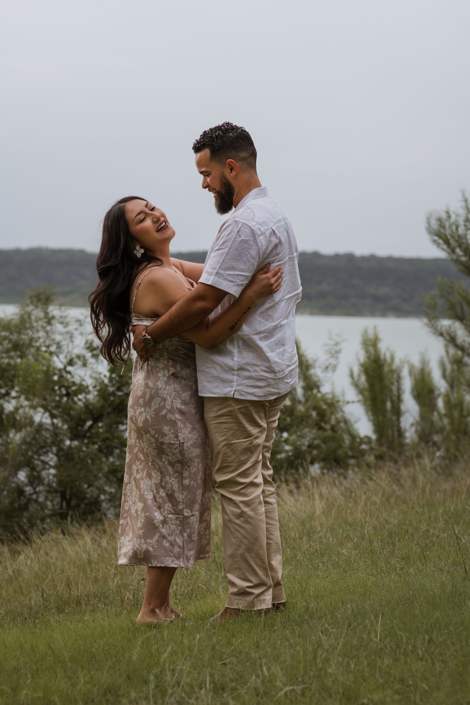 Couple embracing and laughing together on a grassy hillside overlooking a lake on a cloudy day.