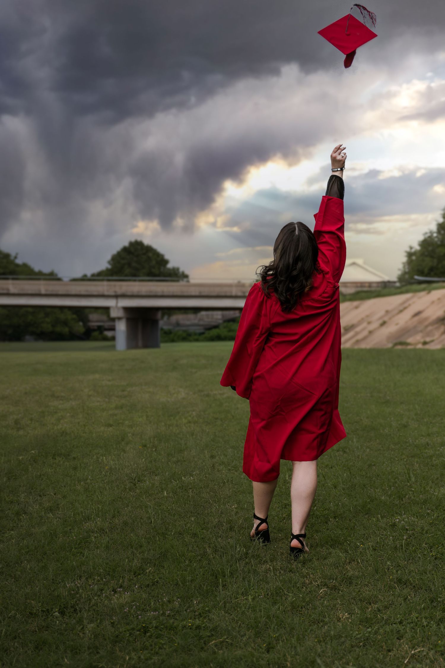 Graduate in red cap and gown tosses mortarboard into stormy sky on green lawn near bridge.