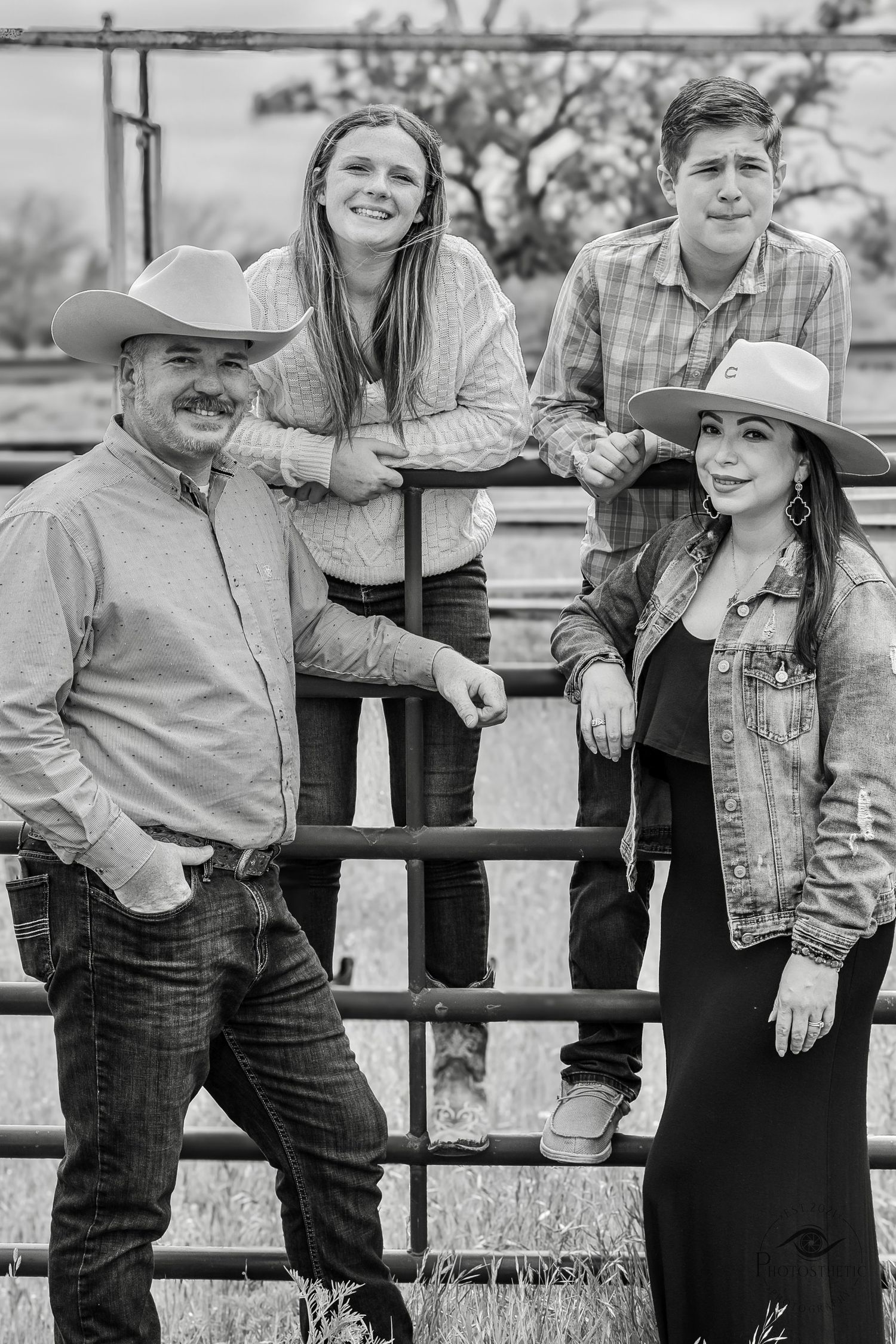 A black and white portrait of a family in western wear posing together against a ranch fence in a rustic outdoor setting.