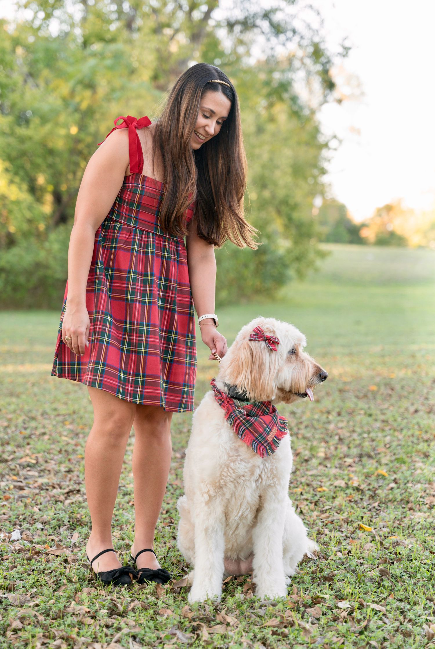 Woman in a red plaid dress standing outdoors in a park with her dog wearing a matching plaid bandana.