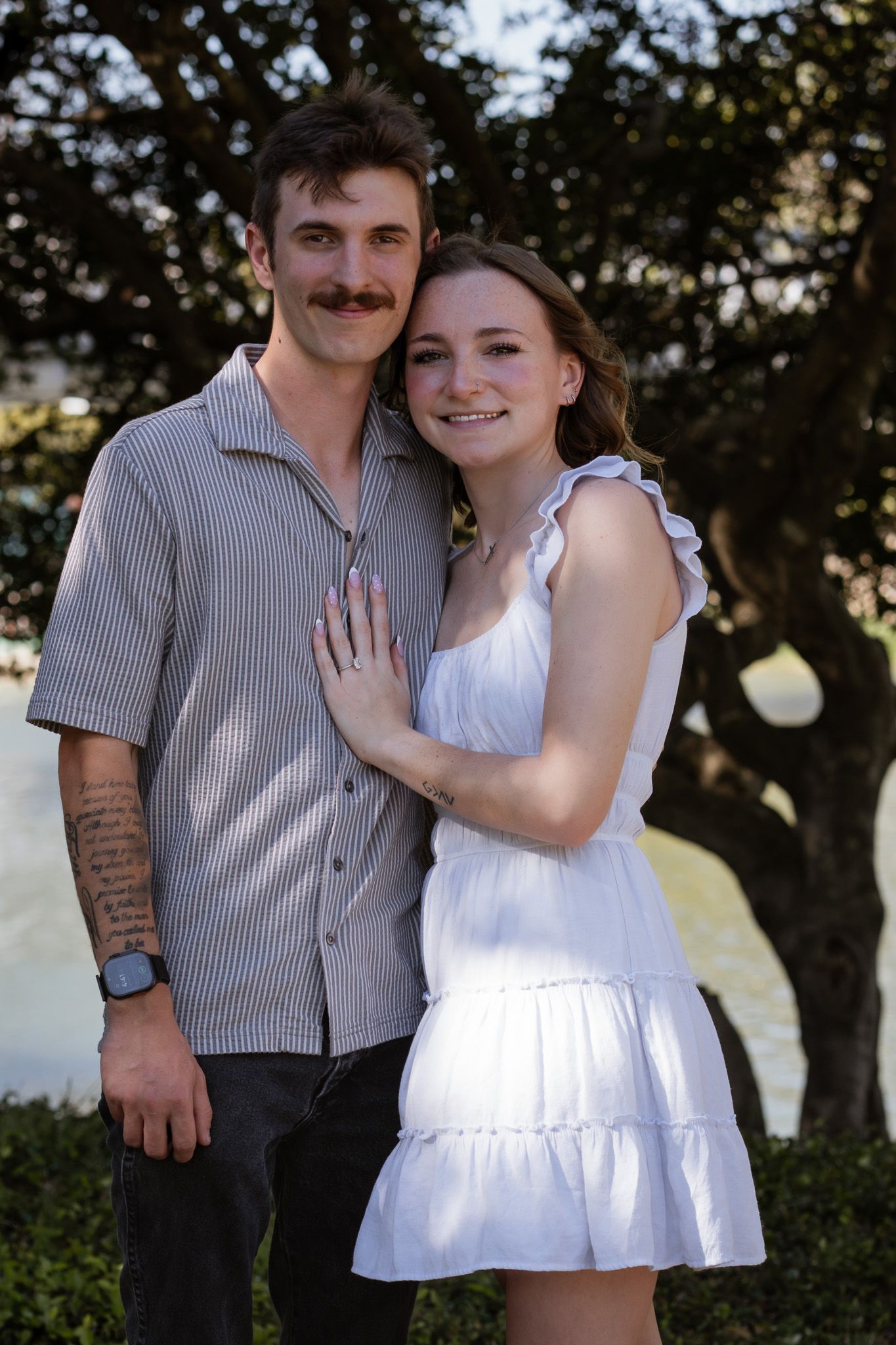 Smiling couple posing outdoors, woman in white dress leaning on man in striped shirt with tattoos, trees in background.
