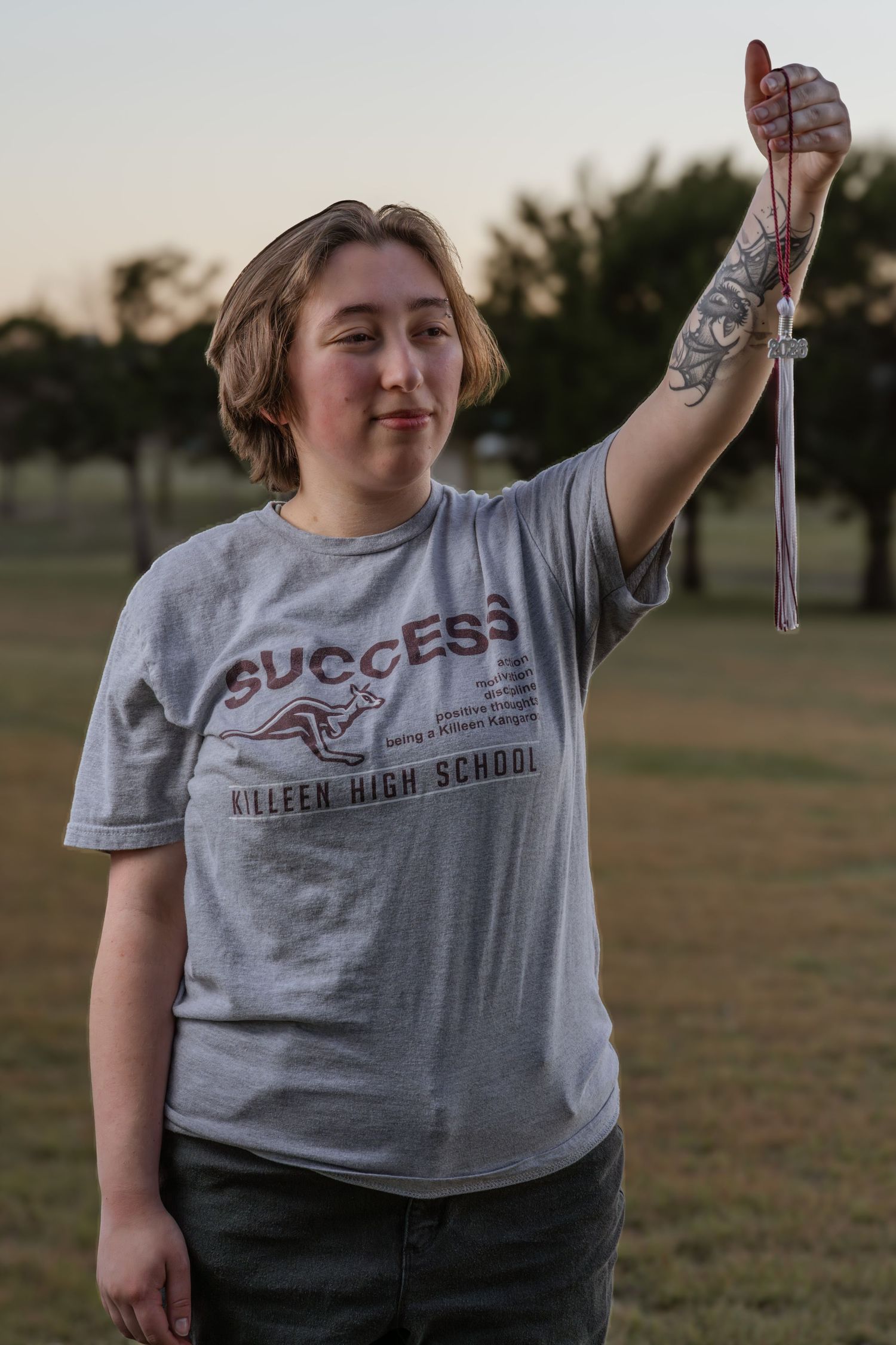 Young woman in Killeen High School Success t-shirt holds up graduation tassel outdoors at dusk.
