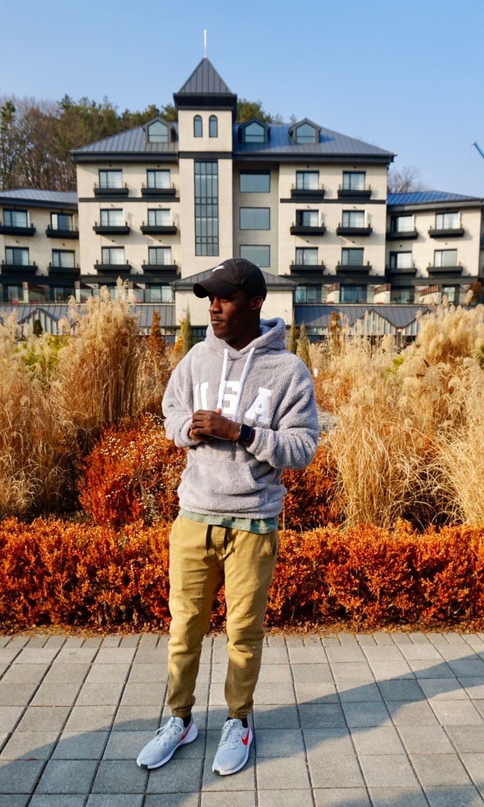 A person in a grey hoodie and khaki pants stands in front of an upscale apartment building with autumn landscaping.