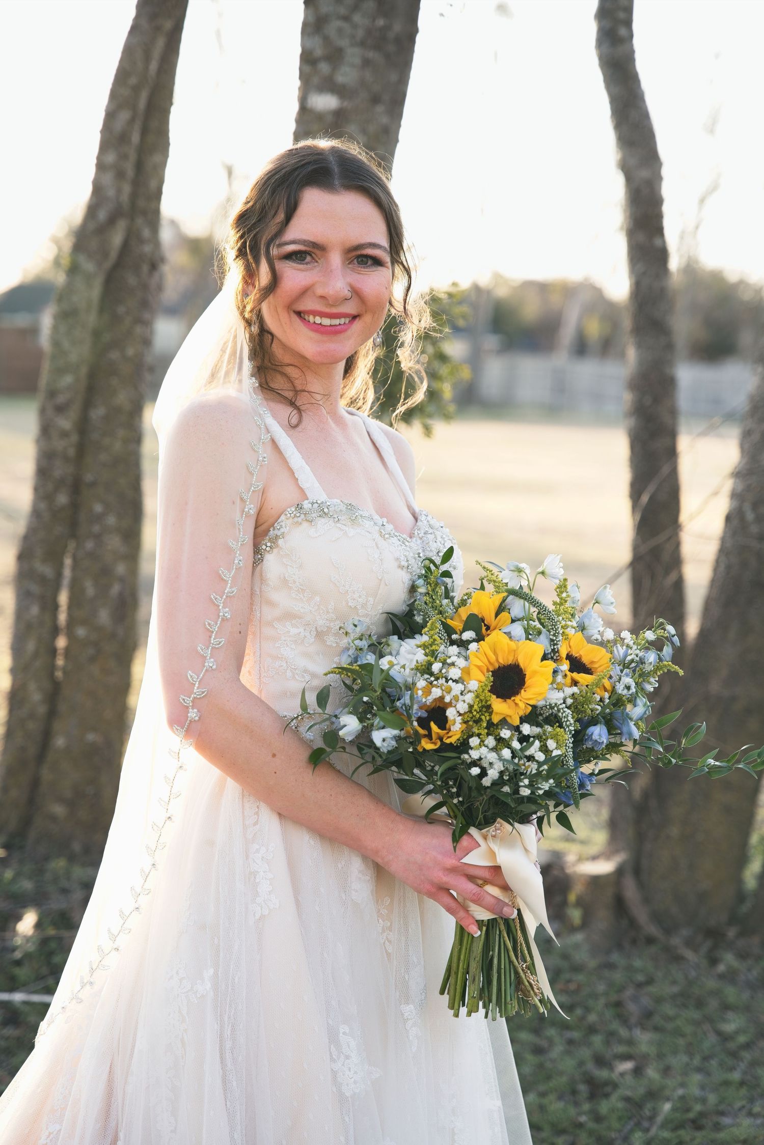 Bride in a lace wedding dress holding a sunflower bouquet outdoors, smiling during a natural light wedding portrait.
