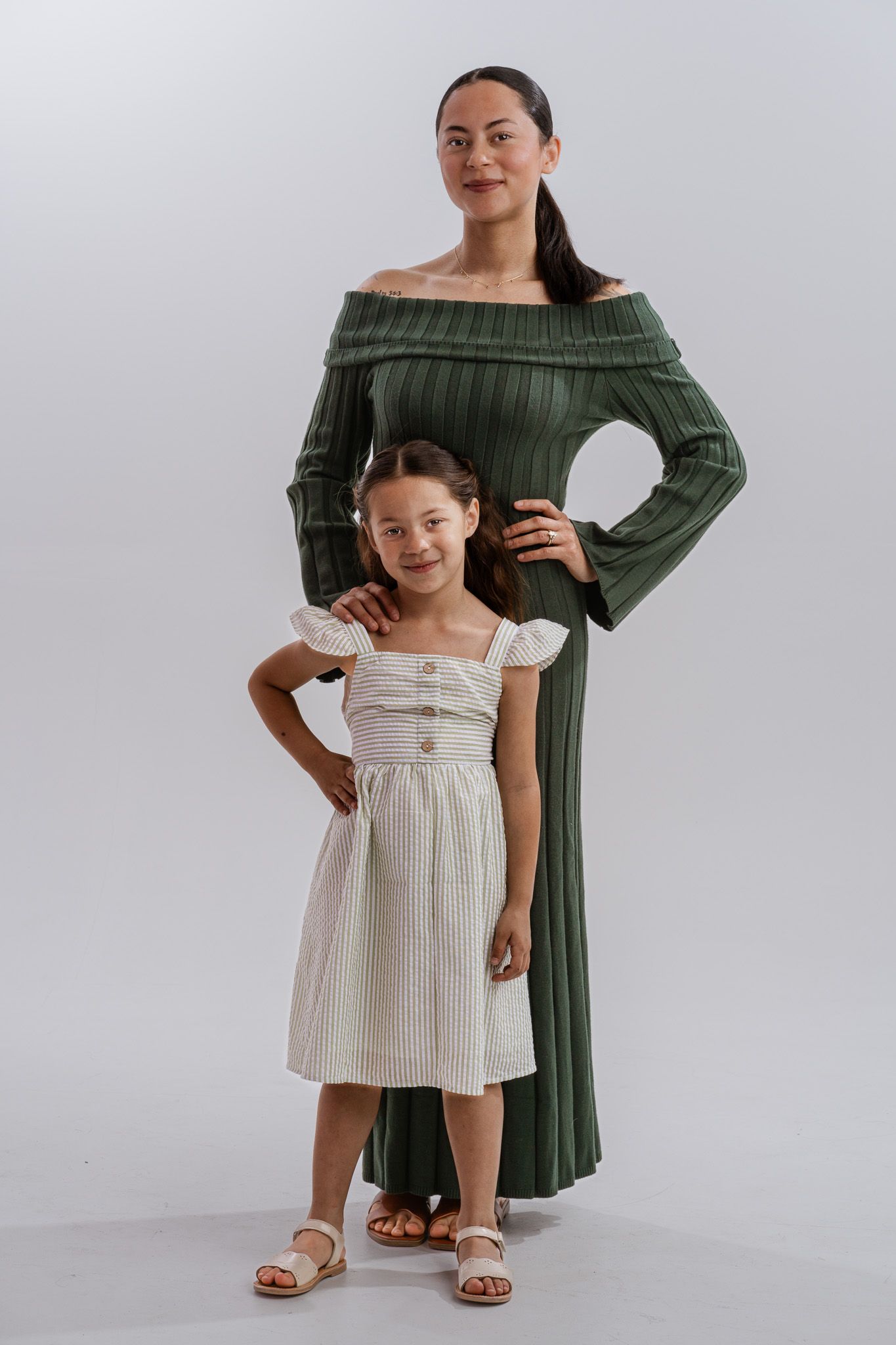 Woman in green off-shoulder dress poses with young girl in white striped midi dress against white background.