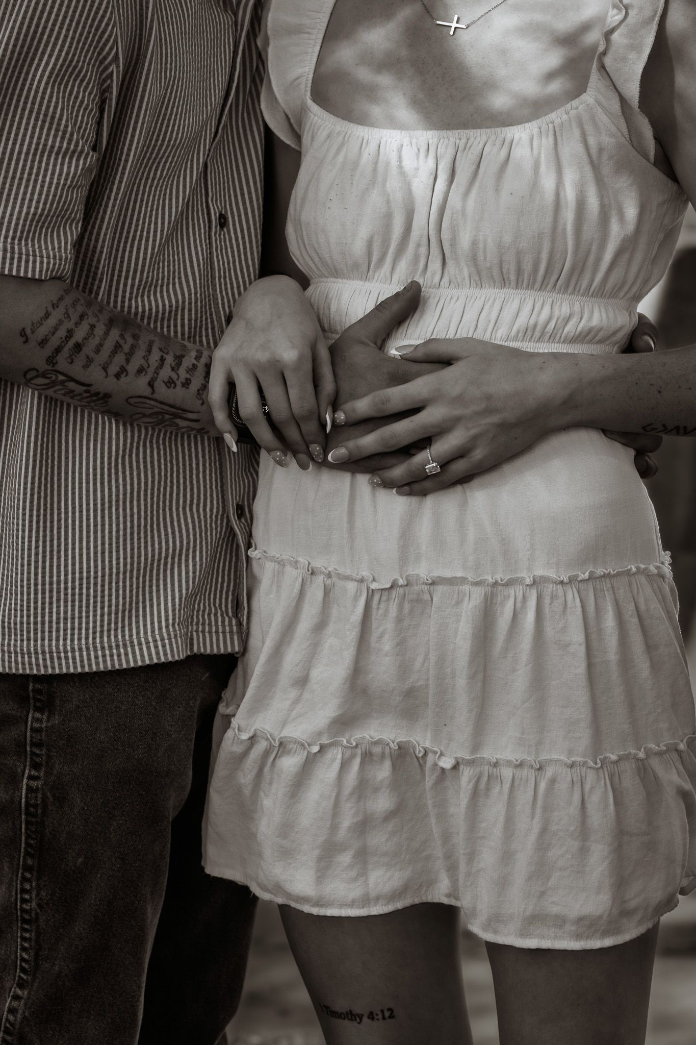 Black and white photo of a couple embracing, woman in white tiered dress with cross necklace, man in striped shirt.
