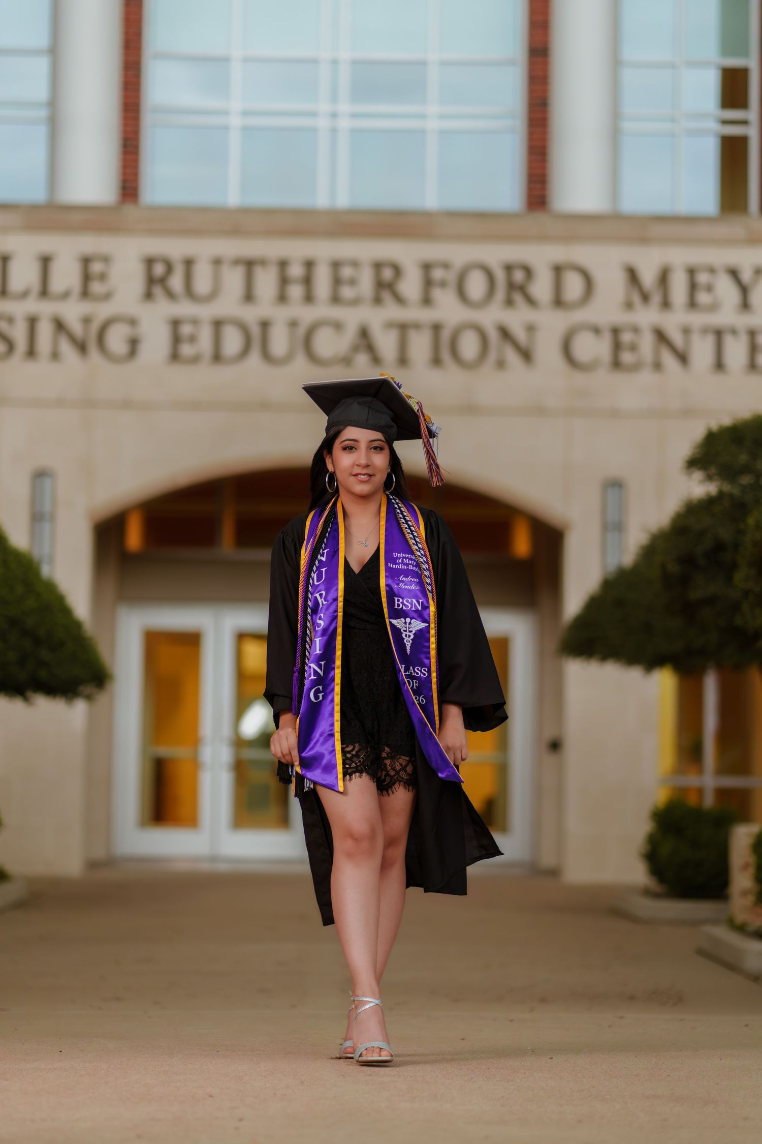 A smiling graduate in black cap and gown with purple stole walks in front of the Rutherford Meyer Nursing Education Center.