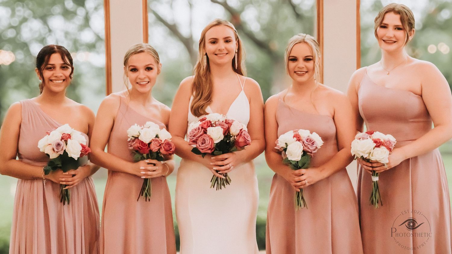 A row of bridesmaids in blush pink dresses stand together holding white floral bouquets at an outdoor wedding.