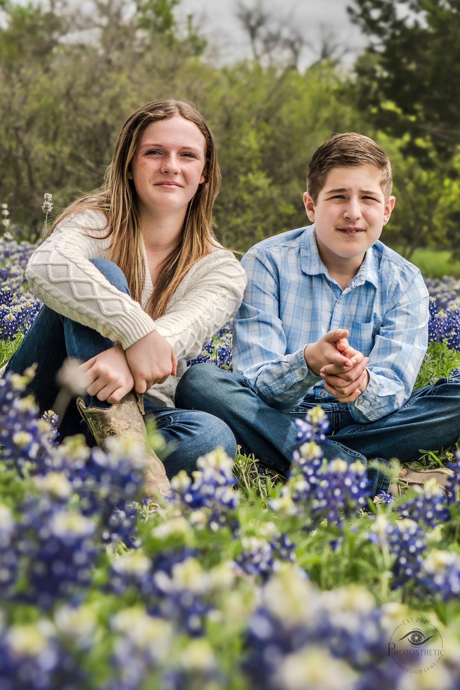 Two siblings sitting in a bluebonnet field smiling during a spring portrait session.