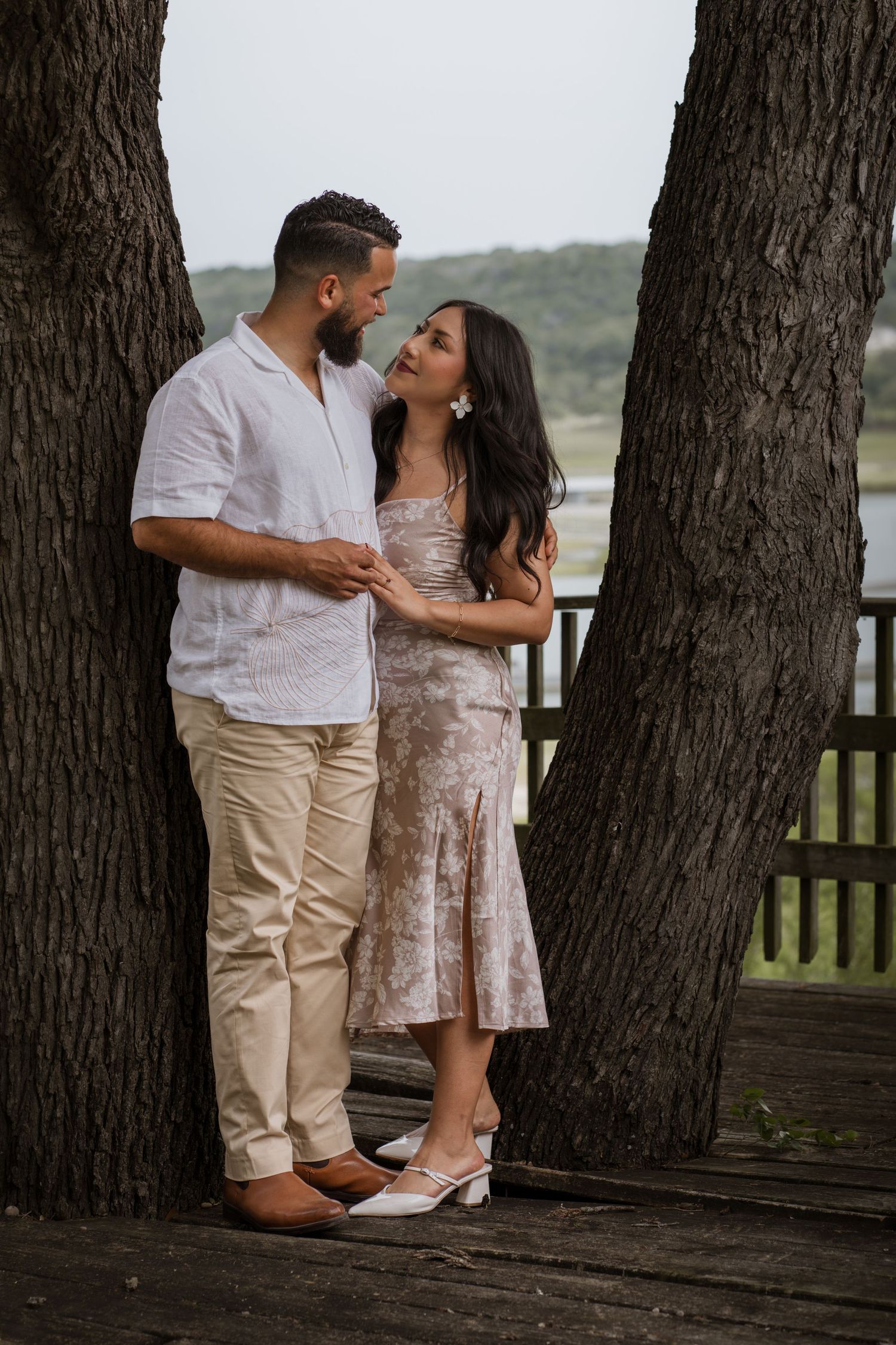 Couple in elegant attire standing between two trees on a wooden deck, sharing a romantic moment outdoors.