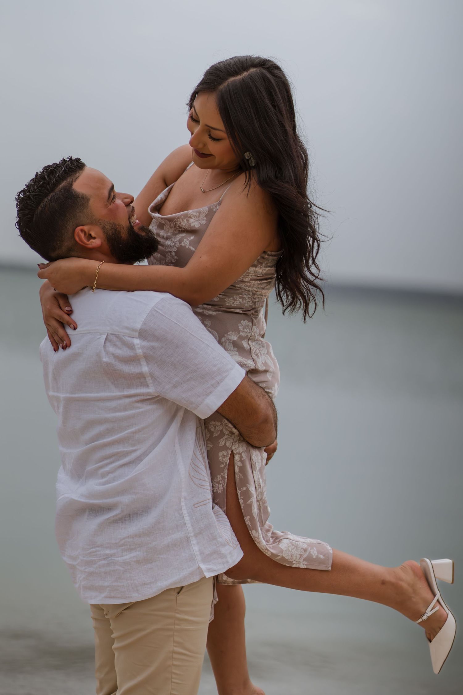 Man lifting woman in floral dress near ocean shore in romantic couples beach photo session.